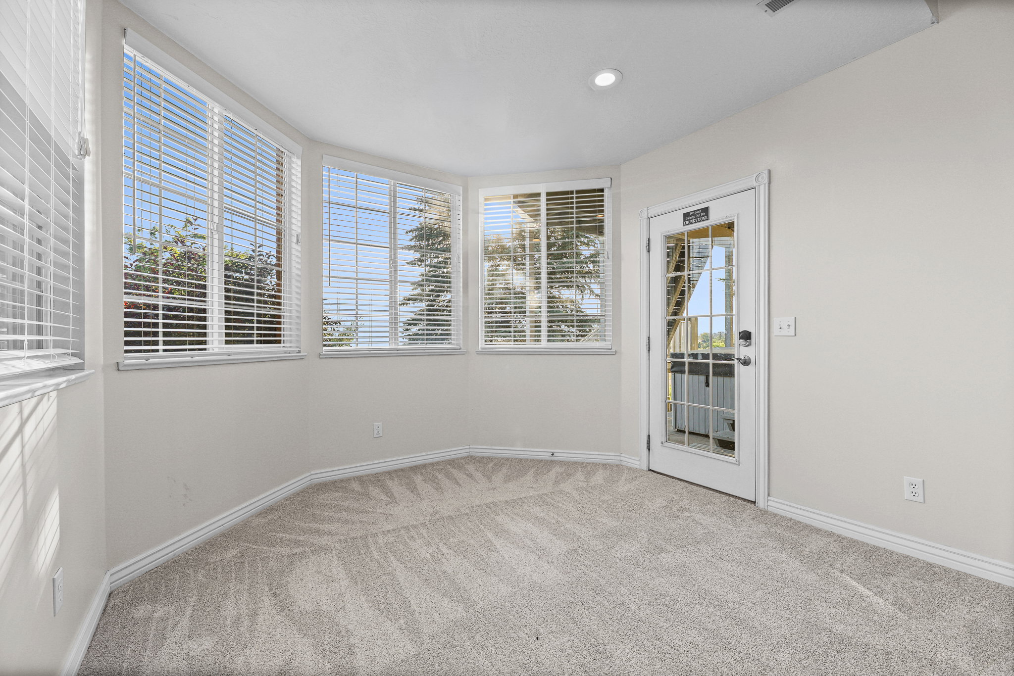 Carpeted room with a bay window design featuring multiple windows with blinds, a recessed ceiling light, and a white door with glass panels