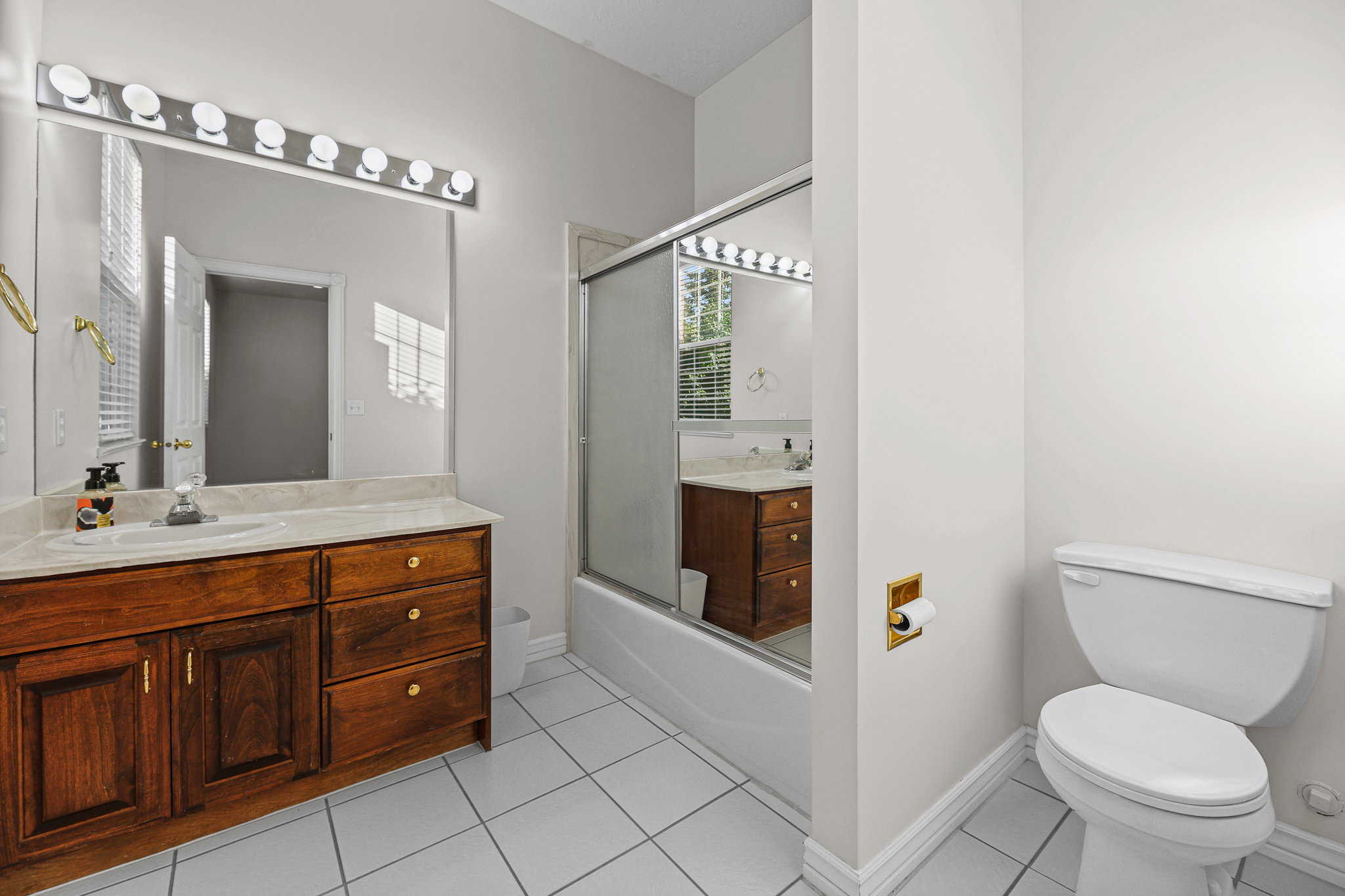 Bathroom featuring a rich wood-finish vanity with an integrated sink, a large wall-mounted mirror, a modern vanity light fixture, a shower-tub combination with a sliding glass door, and white square tile flooring