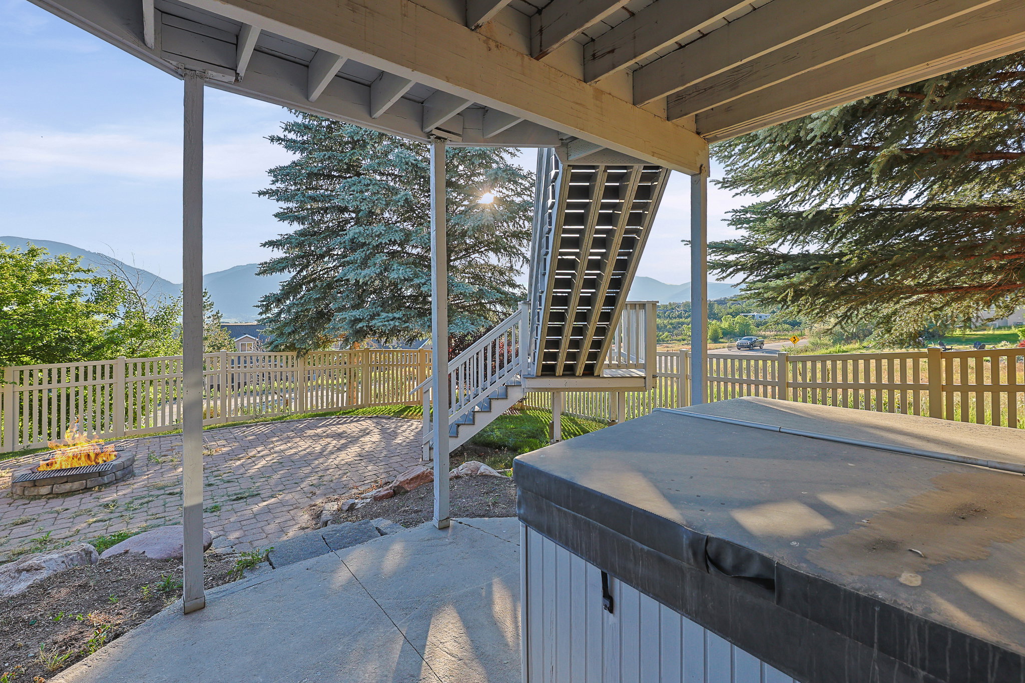 Covered patio area featuring a built-in fire pit, paved patio, wood picket fencing, and mature trees