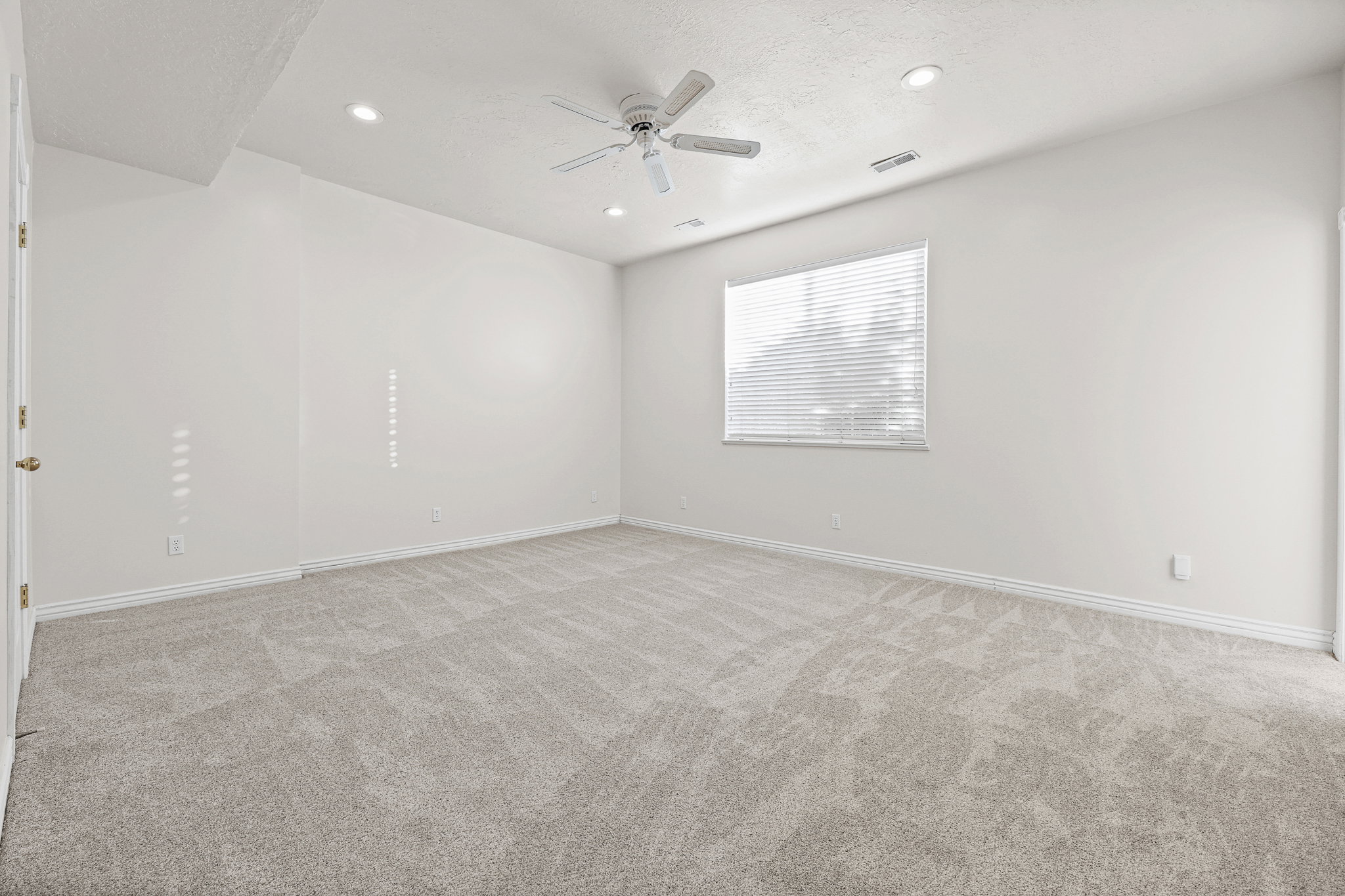 Spacious carpeted room featuring a ceiling fan, recessed lighting, and natural light from a window with blinds