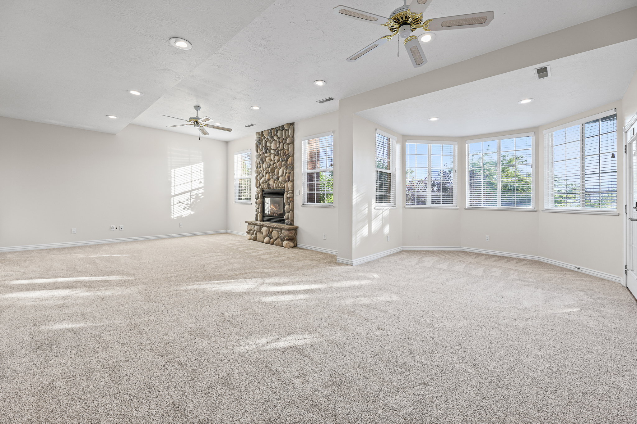 Spacious room featuring a floor-to-ceiling river stone fireplace, bay windows, recessed lighting, and ceiling fans