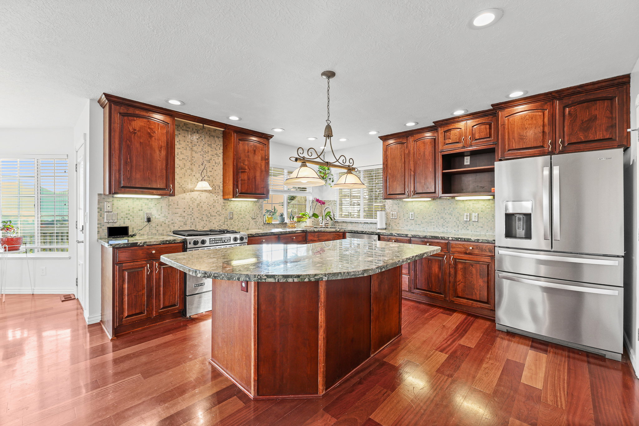 Spacious kitchen featuring rich wood cabinetry, stainless steel appliances, a large central island with a stone countertop, coordinating stone backsplash, and wood-finish flooring