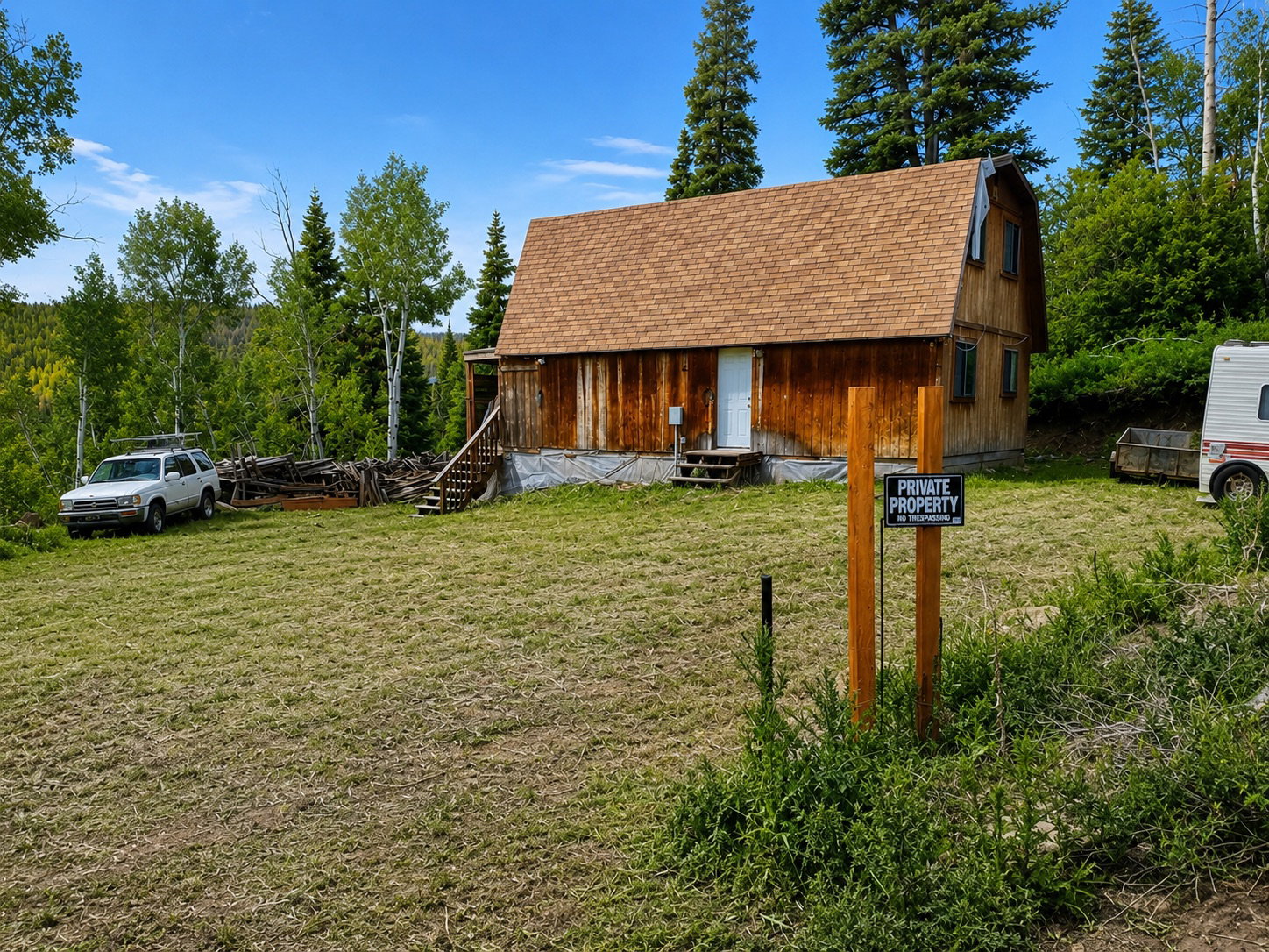 Wood-sided structure featuring a gambrel roof and multiple windows, surrounded by a natural landscape of mature trees and a grassy area