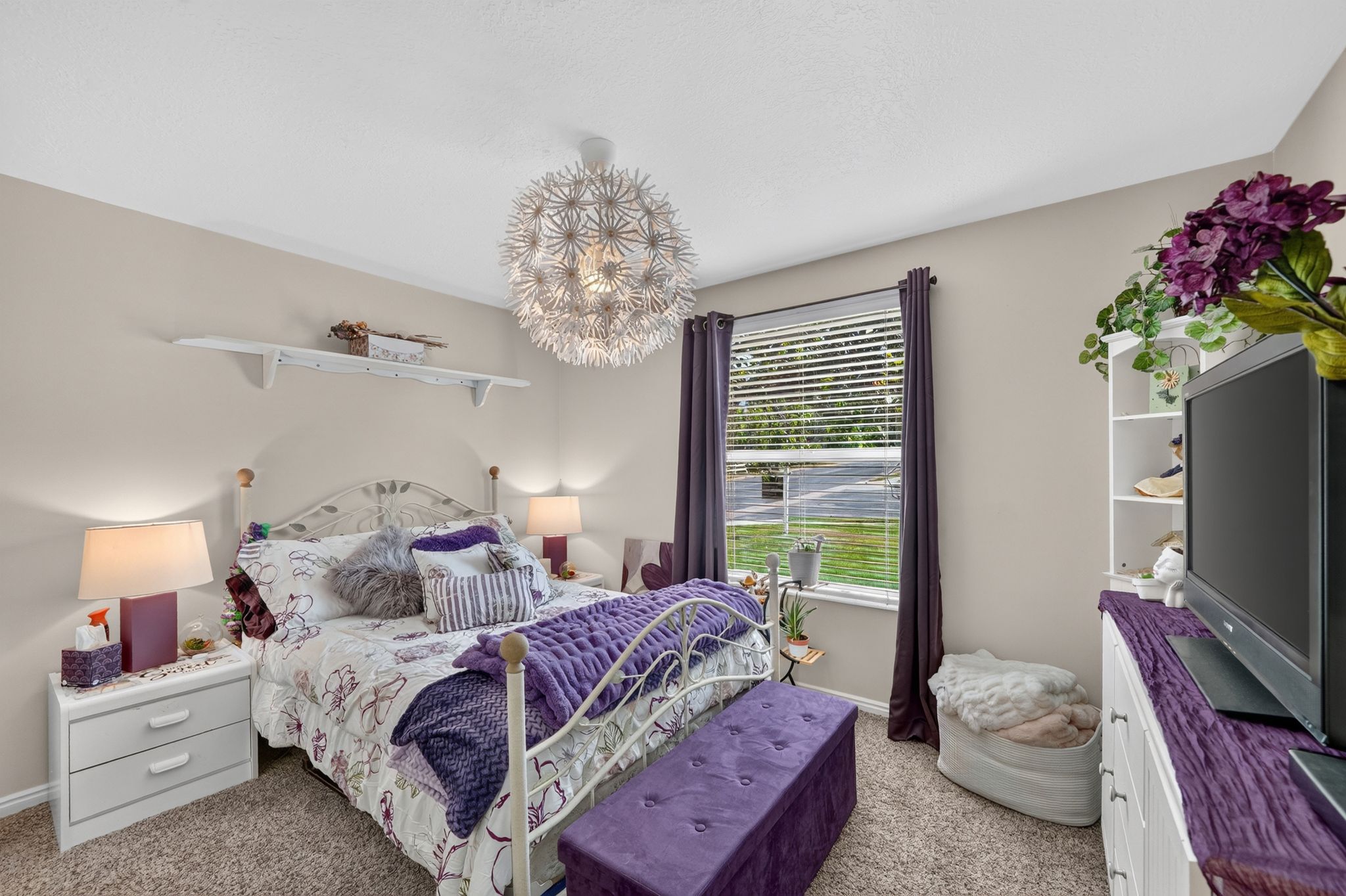 Carpeted room featuring a large window with blinds and dark curtains, a built-in shelving unit, a ceiling-mounted light fixture, and neutral painted walls