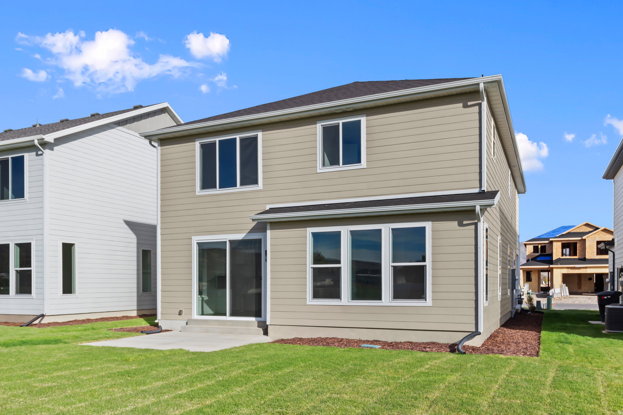 Rear exterior featuring beige horizontal siding, a sliding glass door, multiple large windows, a concrete patio, and a grass lawn