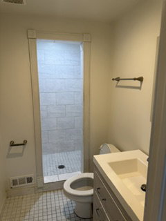 Bathroom featuring a walk-in shower with tile surround, a single vanity with an integrated sink, a toilet, and grid-pattern floor tiling