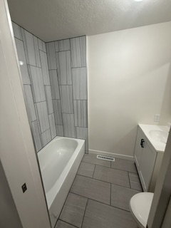Bathroom featuring a white bathtub with tile surround, a single vanity with storage, a toilet, and tile flooring