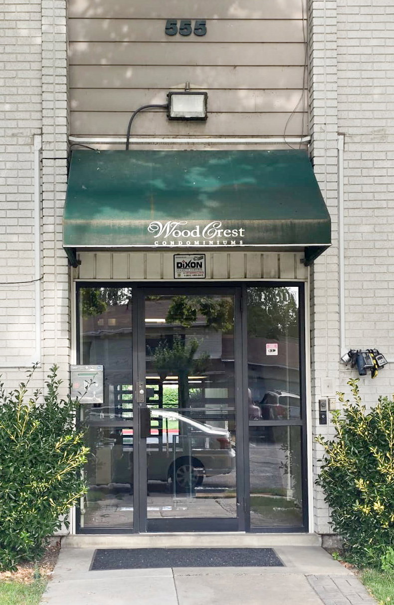 Building entrance featuring a prominent green awning, glass double doors, a white brick facade, and landscaping