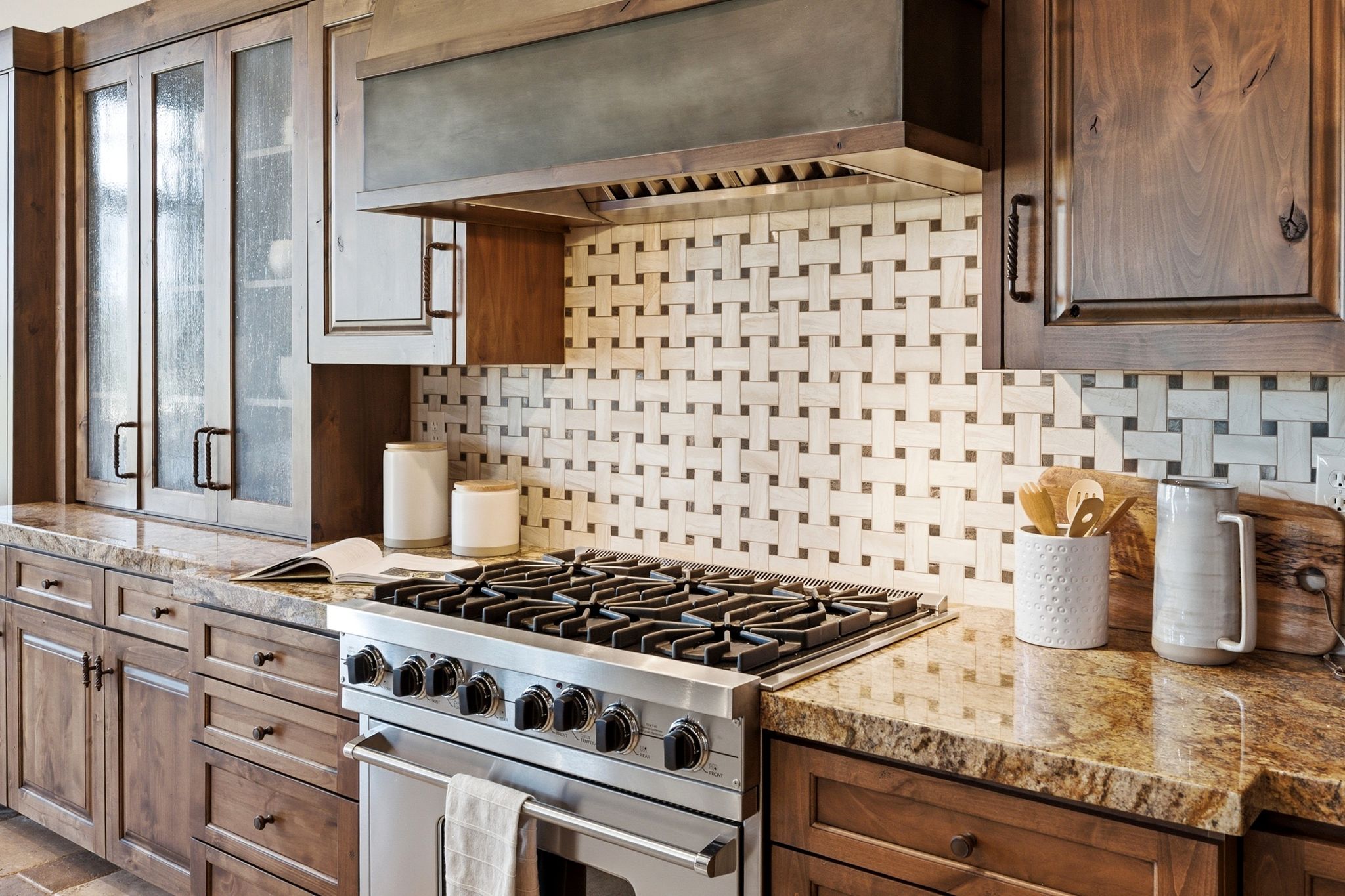 Stainless steel range with multiple burners, stone countertops, custom wooden cabinetry, decorative tile backsplash, and a metal range hood