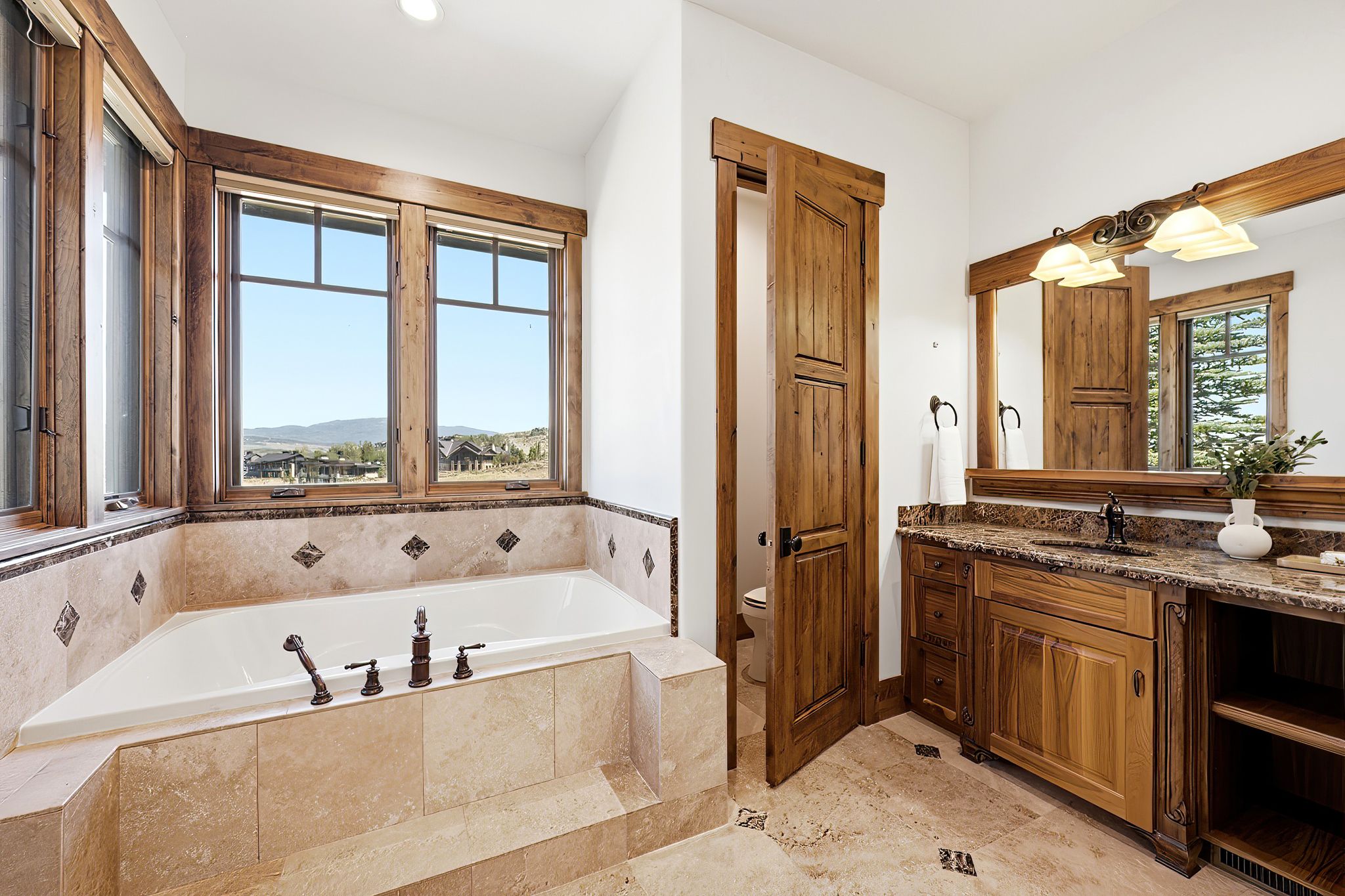 Soaking tub with bronze fixtures, surrounded by tiled decking and large wood-framed windows