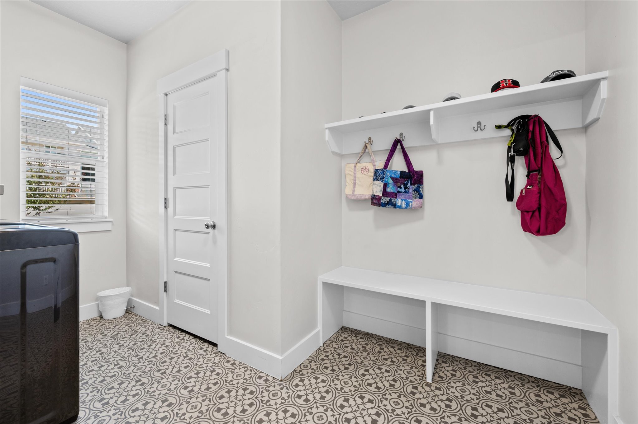 Utility room featuring patterned flooring, a window with blinds, a built-in bench, and a wall-mounted shelf with coat hooks