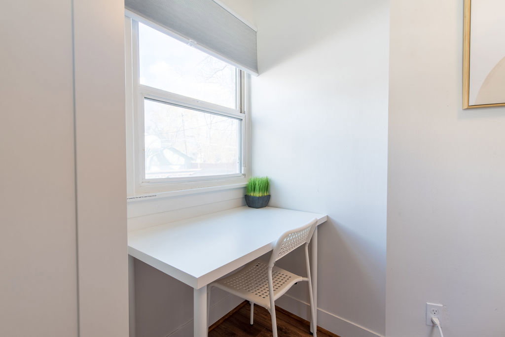 Built-in desk nook featuring a white countertop, window with roller shade, white walls, and wood-finish flooring