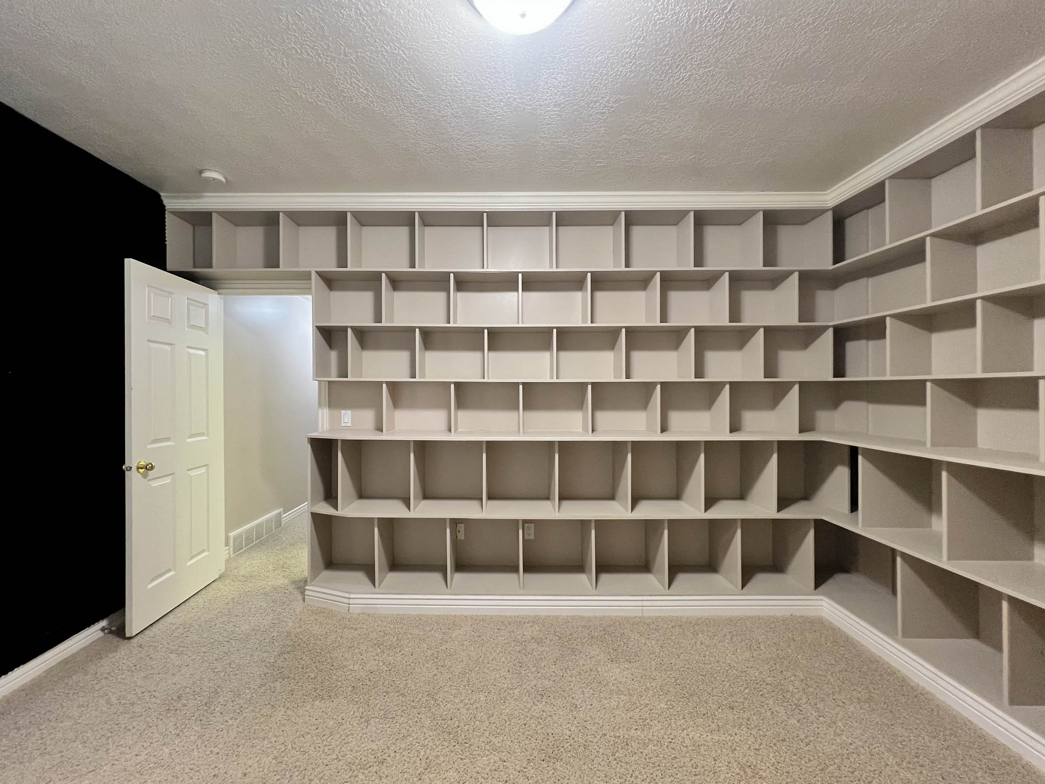Built-in shelving units encompassing multiple walls, crown molding, light-colored carpet flooring, and a flush-mount ceiling light fixture