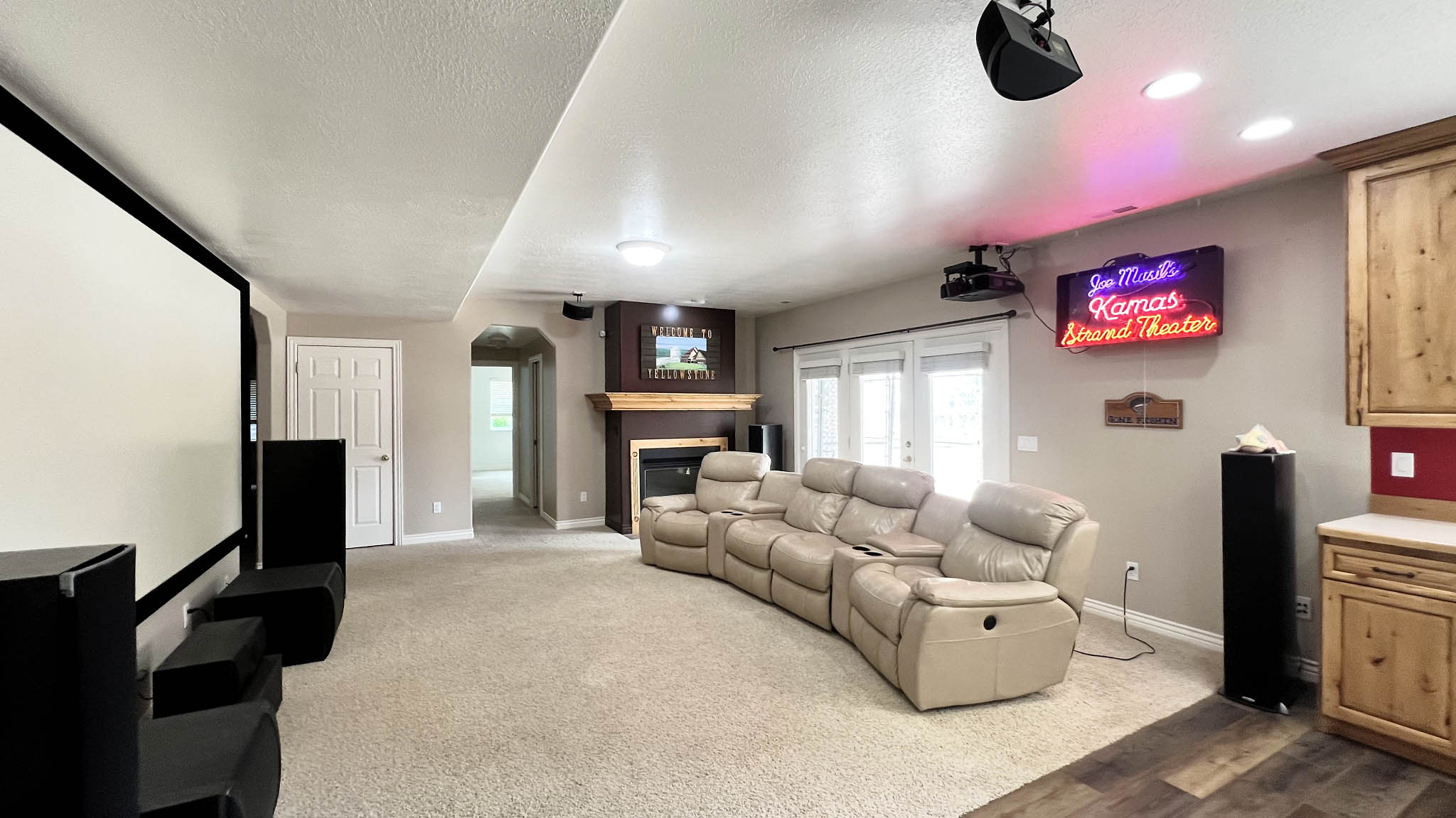 Spacious entertainment room featuring a large projection screen, recessed lighting, carpet flooring, a corner fireplace with wood mantel, and built-in cabinetry with wood-finish flooring