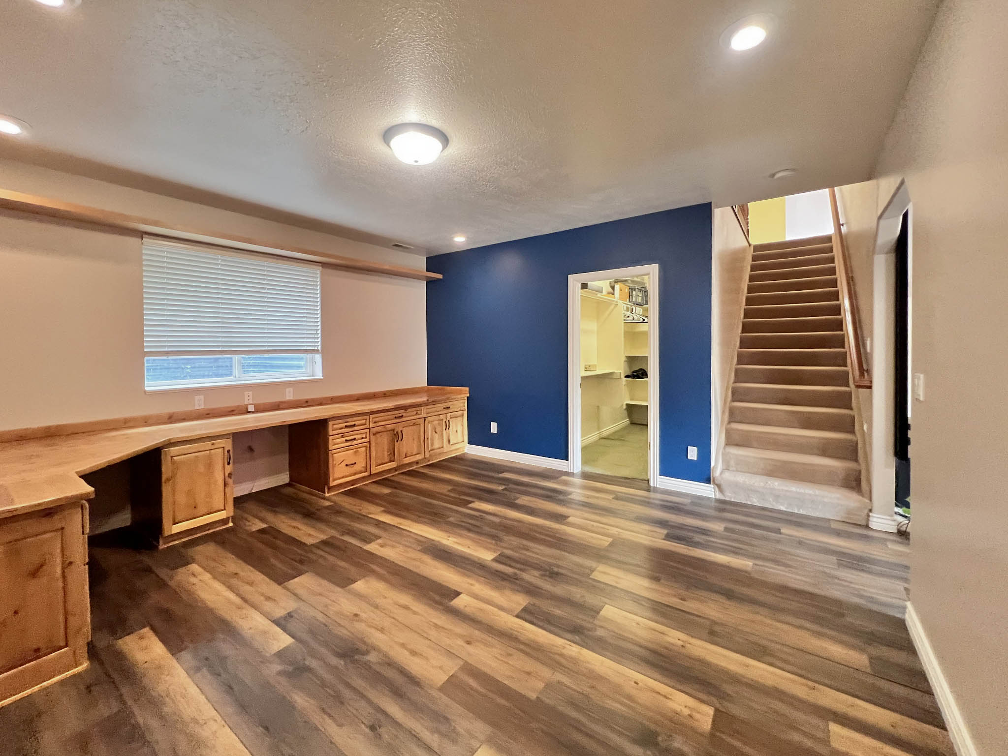 Spacious room featuring wood-finish flooring, a built-in desk with natural wood cabinetry, a window with blinds, and recessed lighting