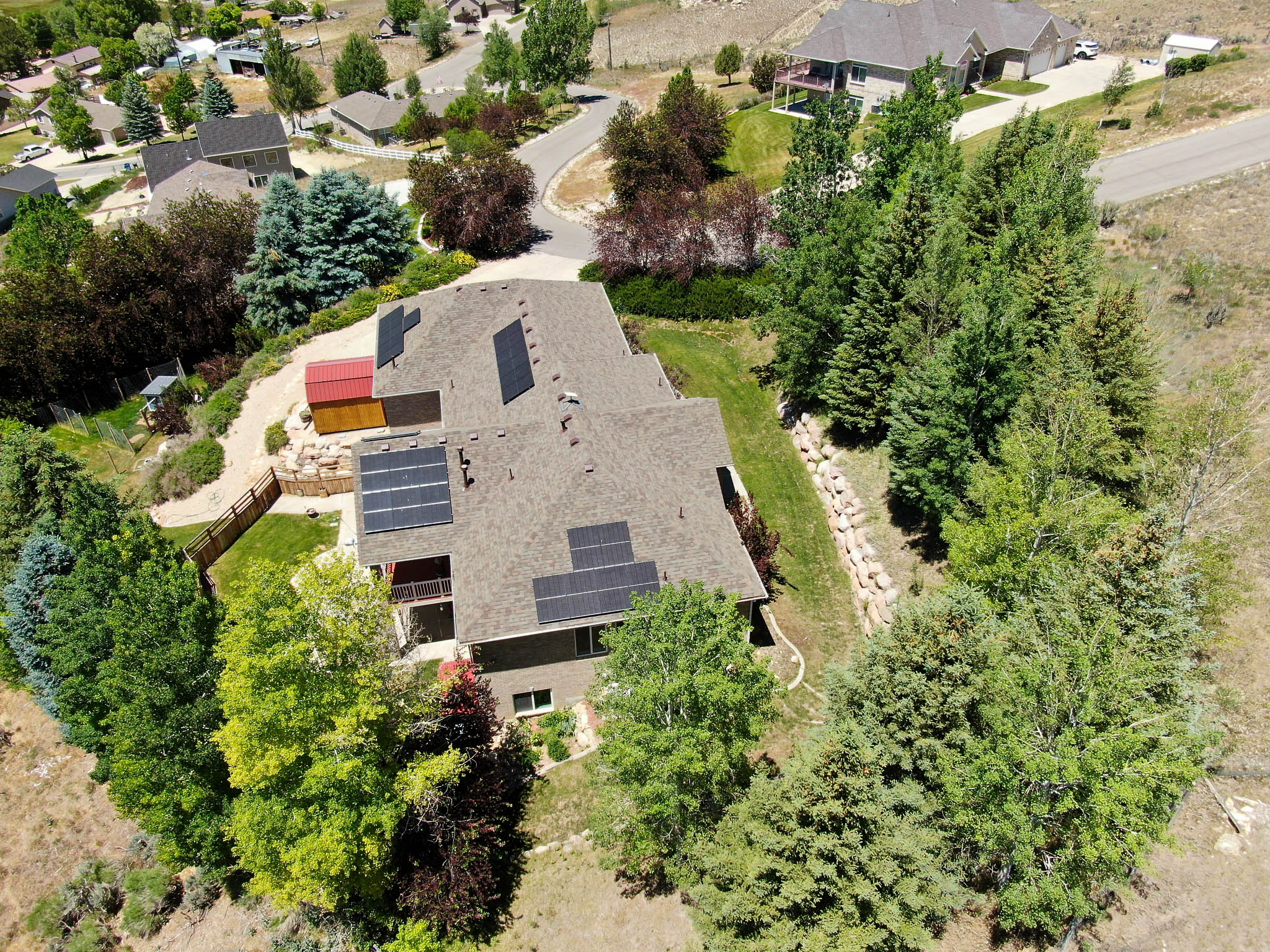 Architectural property featuring a shingle roof with multiple solar panel installations, a ground-level patio, and a terraced backyard with mature trees