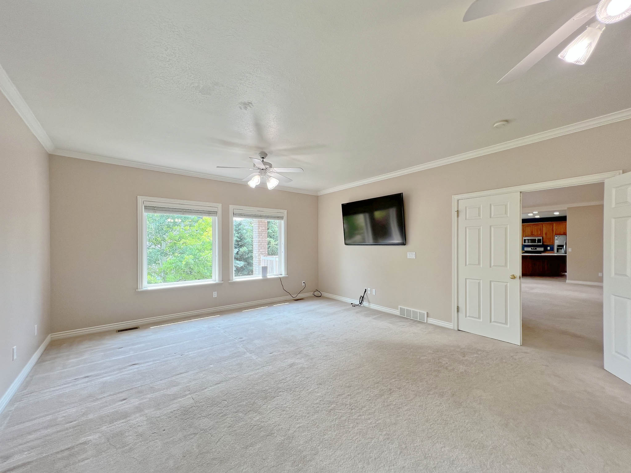 Spacious room featuring neutral carpeting, two large windows, dual ceiling fans, and an open doorway leading to a kitchen area