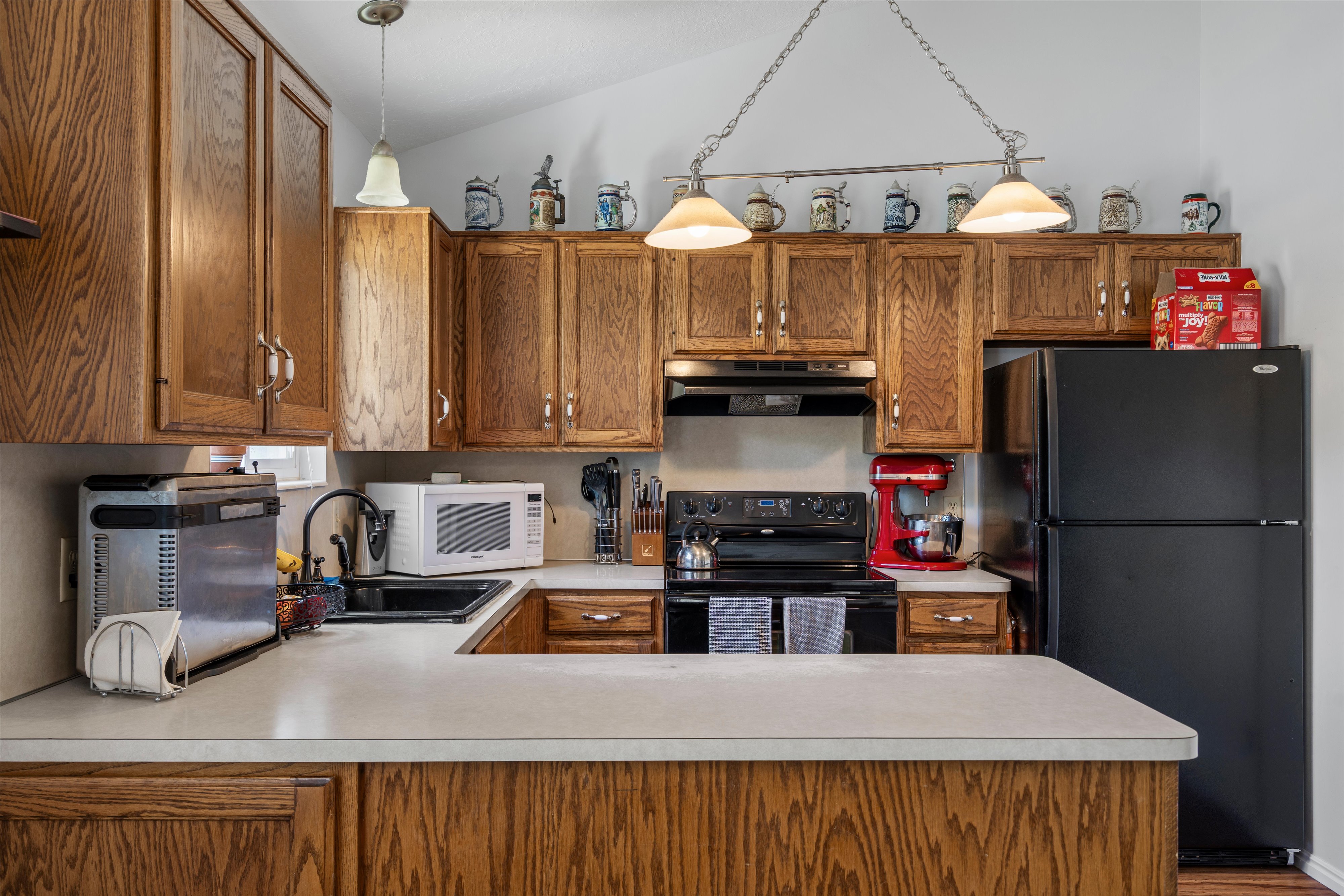 Kitchen featuring wood cabinetry with silver hardware, a black electric range with an overhead vent, a black double-door refrigerator, a black sink, and light-colored countertops