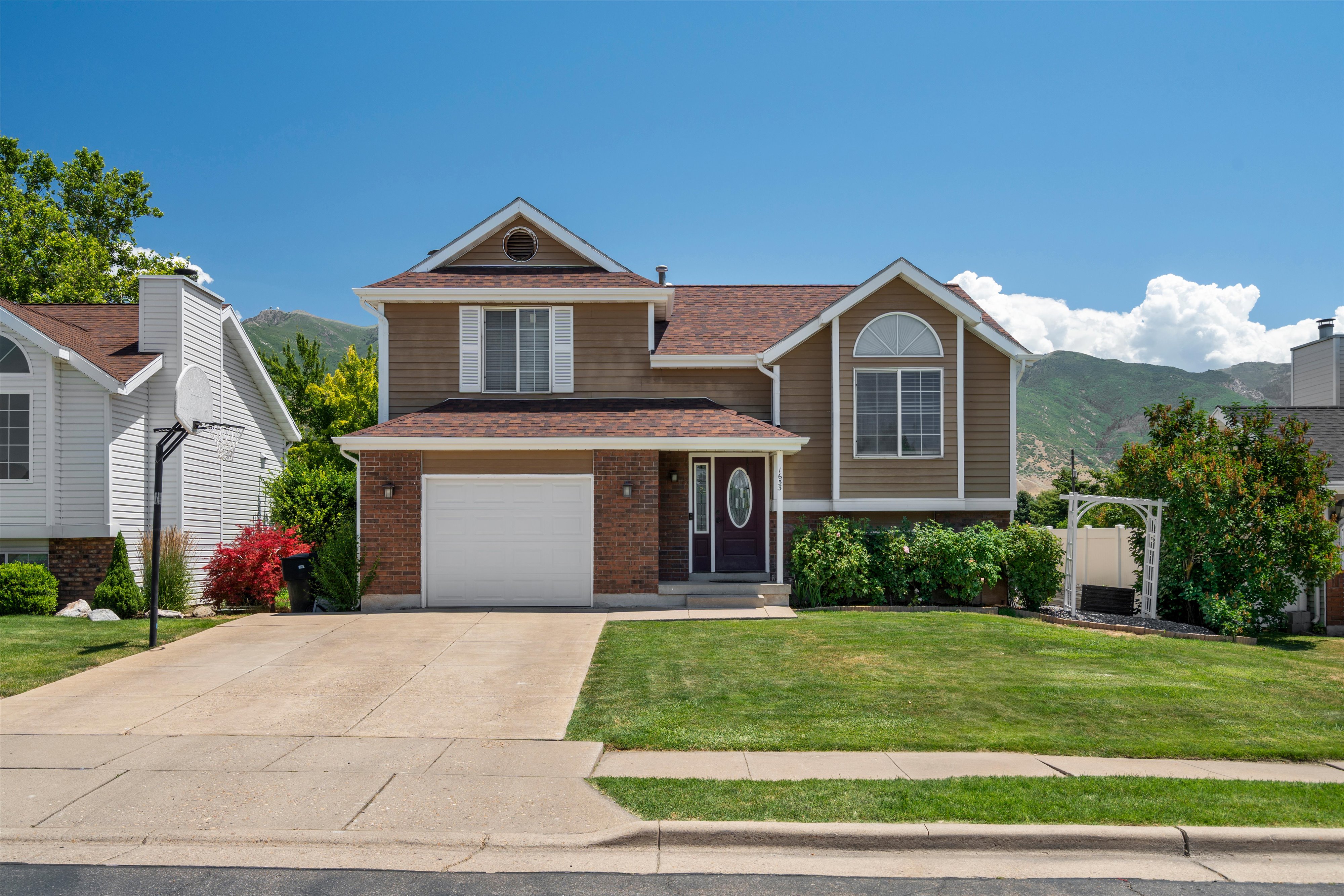 Two-story residence featuring a brown siding and brick exterior, an attached garage, a front lawn, and mature landscaping