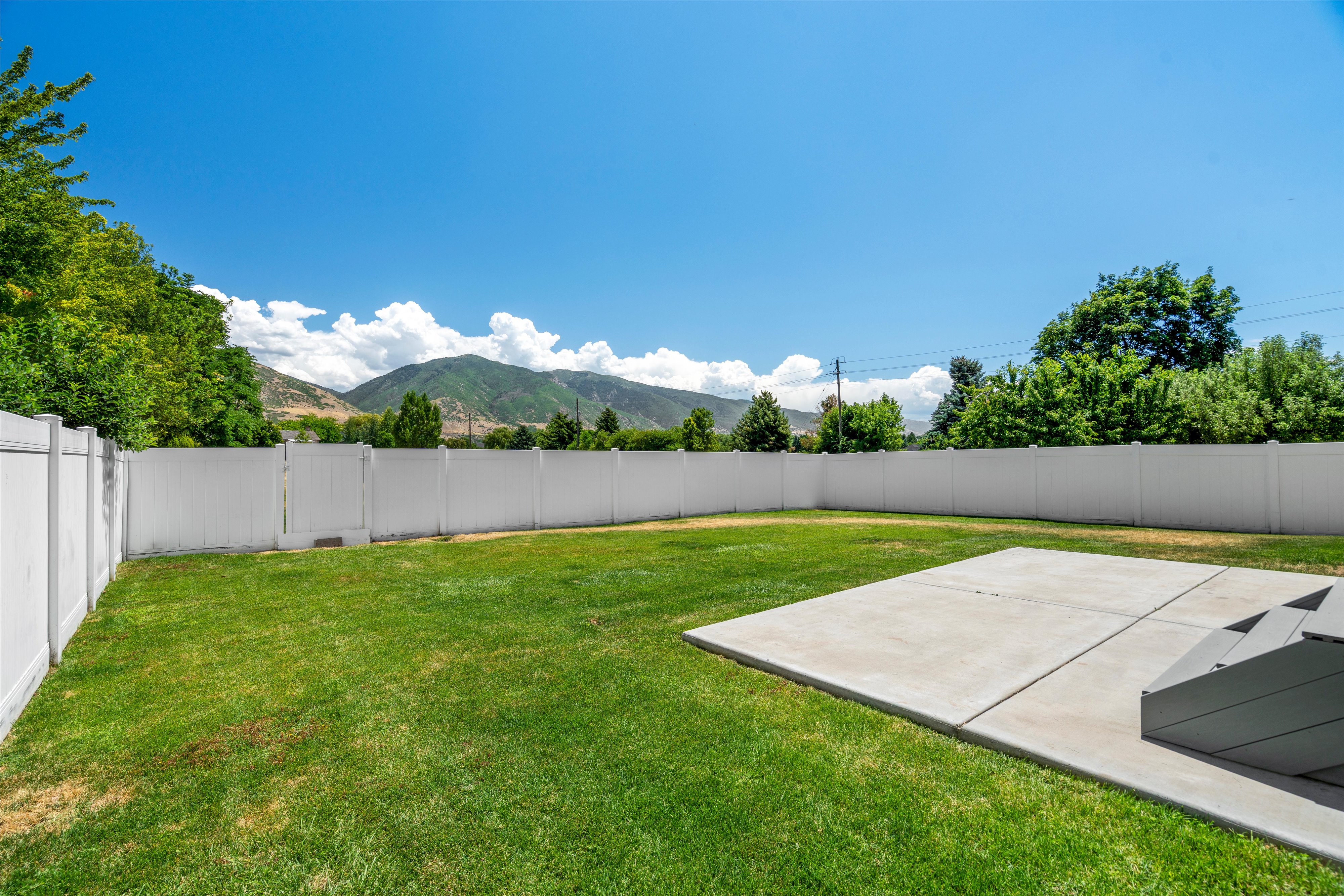 Expansive backyard featuring a lush lawn, white privacy fencing, a concrete patio, and mountain scenery