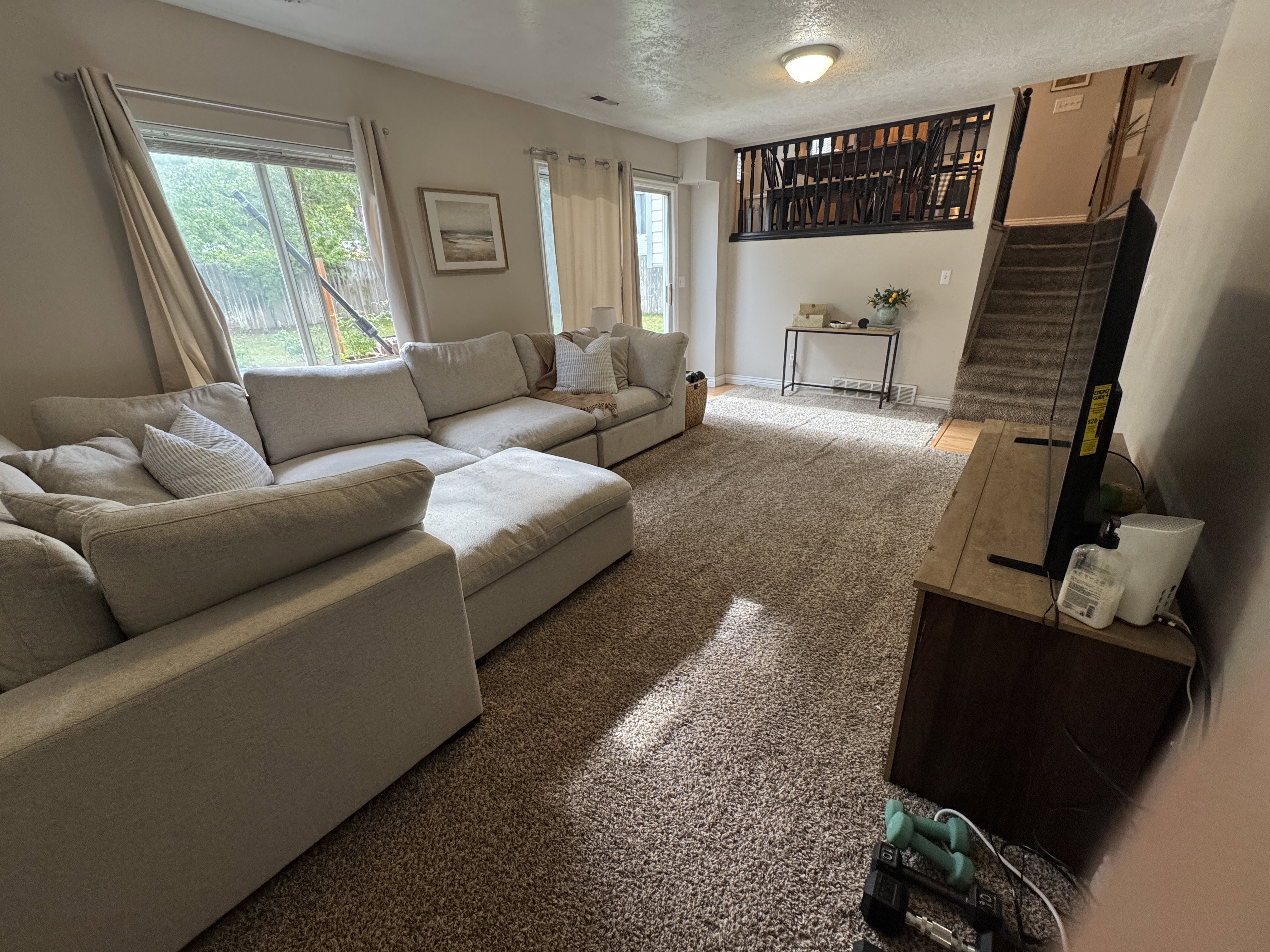 Spacious living area featuring neutral-toned carpeting, ample natural light from multiple windows, and a multi-level layout with a visible staircase and upper-level railing