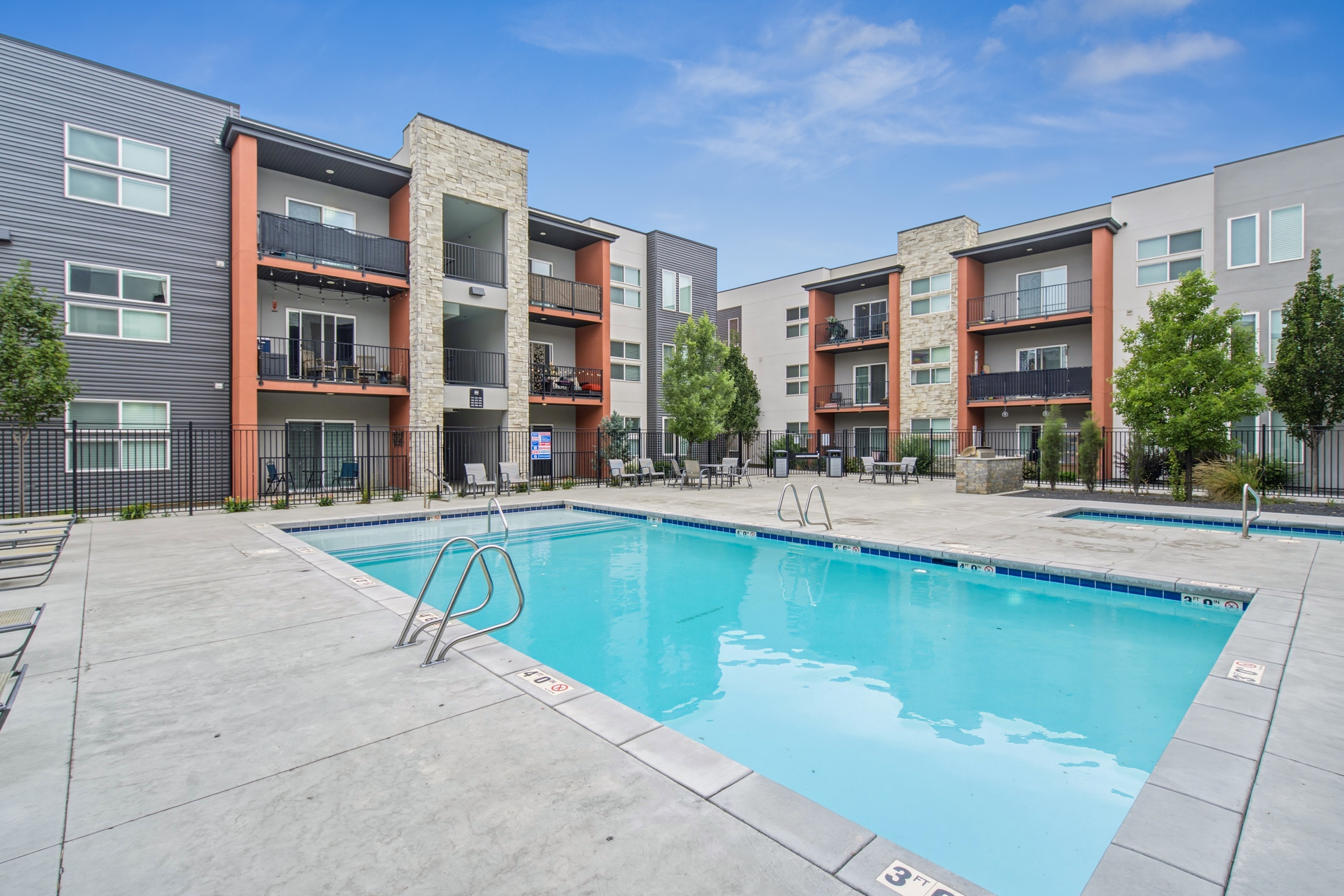 Outdoor swimming pool with concrete decking, surrounded by multi-story buildings featuring balconies and a mix of exterior siding and stone accents