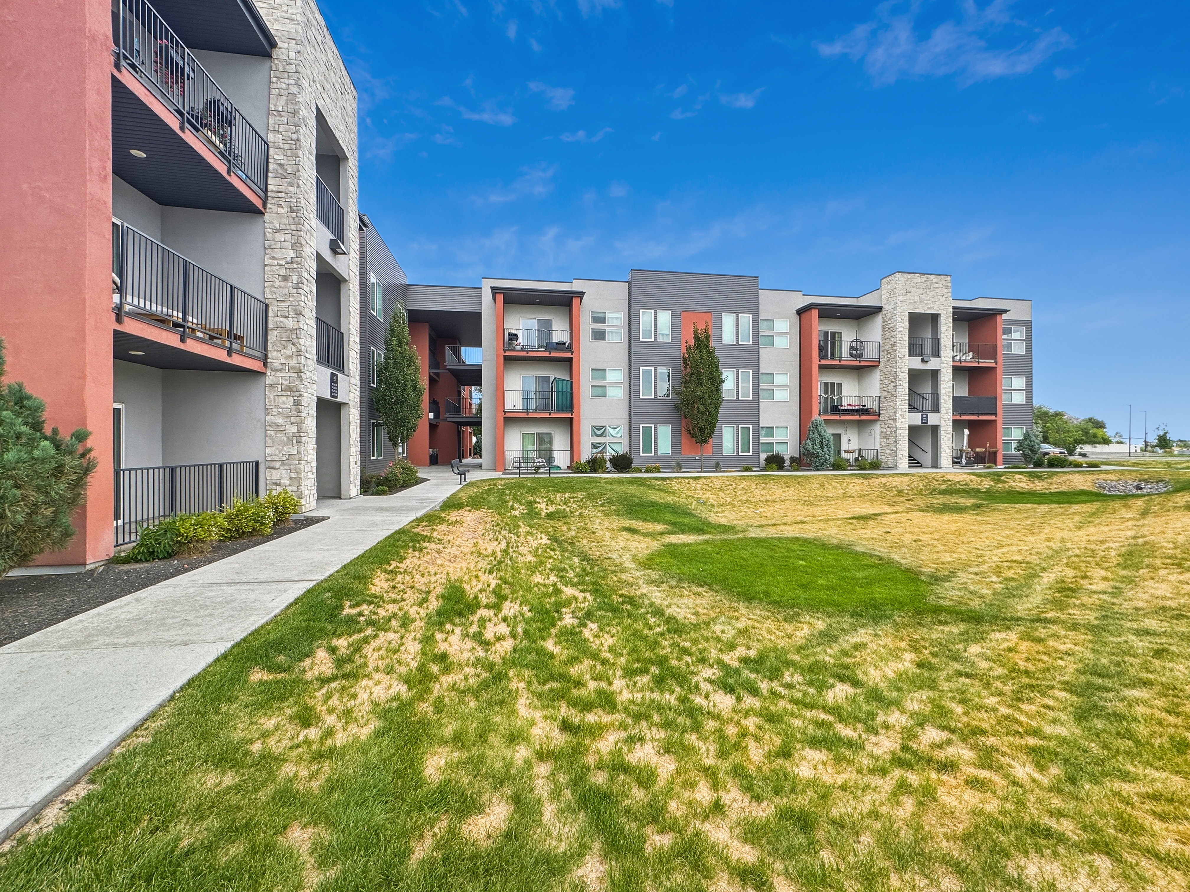 Contemporary multi-story building featuring stone and panel siding, private balconies with metal railings, and a paved pathway alongside a grassy area