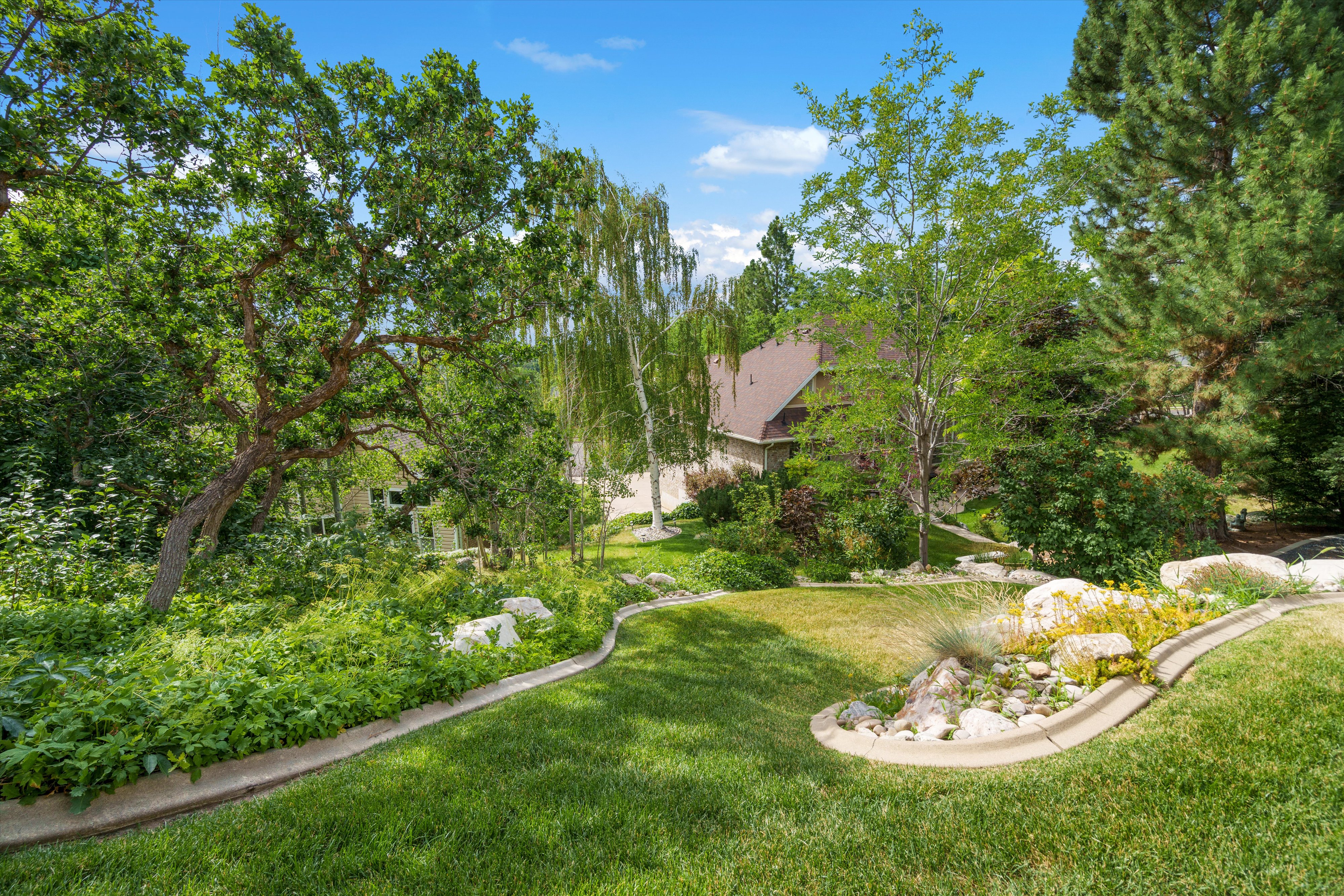 Manicured lawn with a decorative rock garden featuring varied plantings, mature trees, and a visible portion of the property's exterior