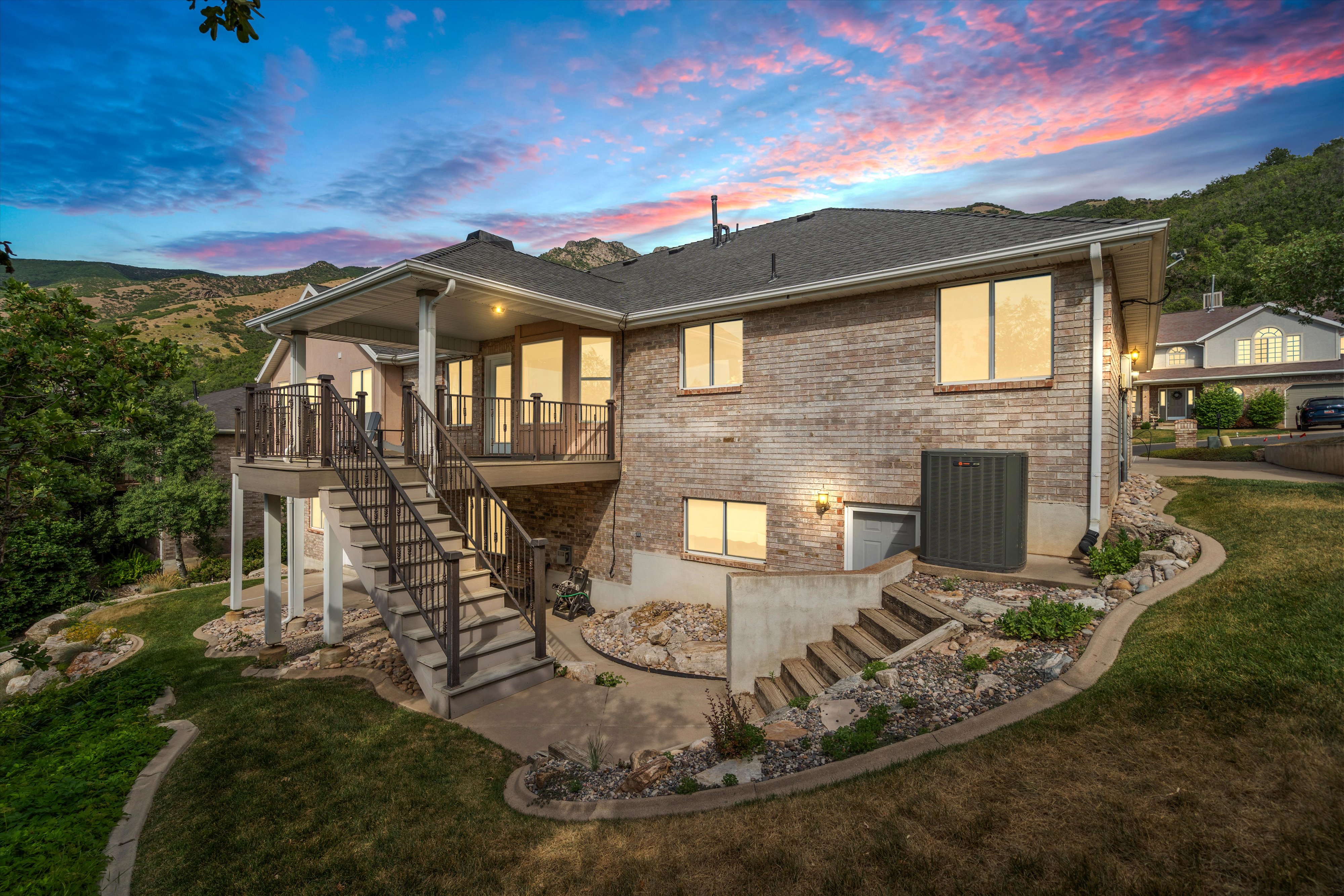 Rear elevation featuring a brick facade, covered deck with dark railings, exterior staircase, and lower-level patio access