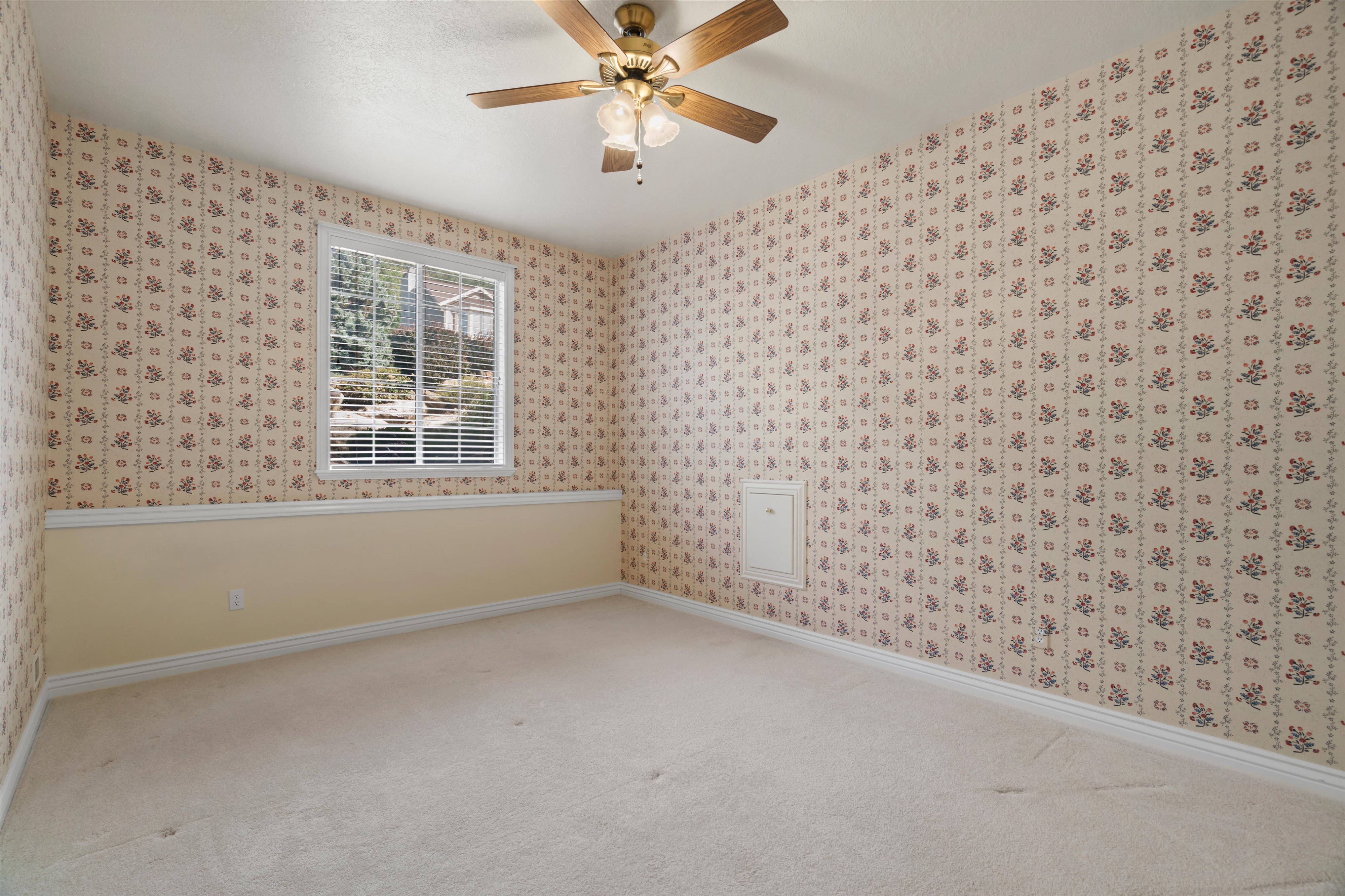 Carpeted interior space featuring patterned wallpaper, a single window with horizontal blinds, a ceiling fan with integrated lighting, and a white access panel
