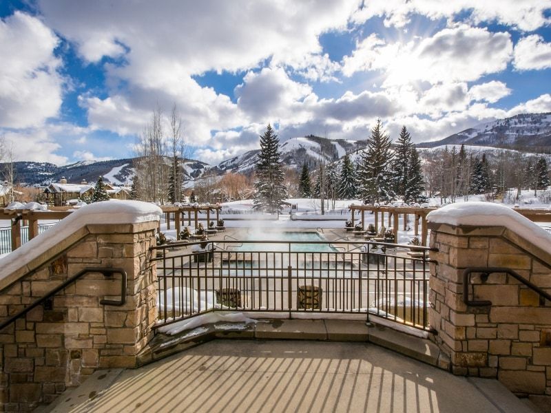 Outdoor pool and spa area with stone and metal railing, surrounded by snow-covered trees and distant mountains