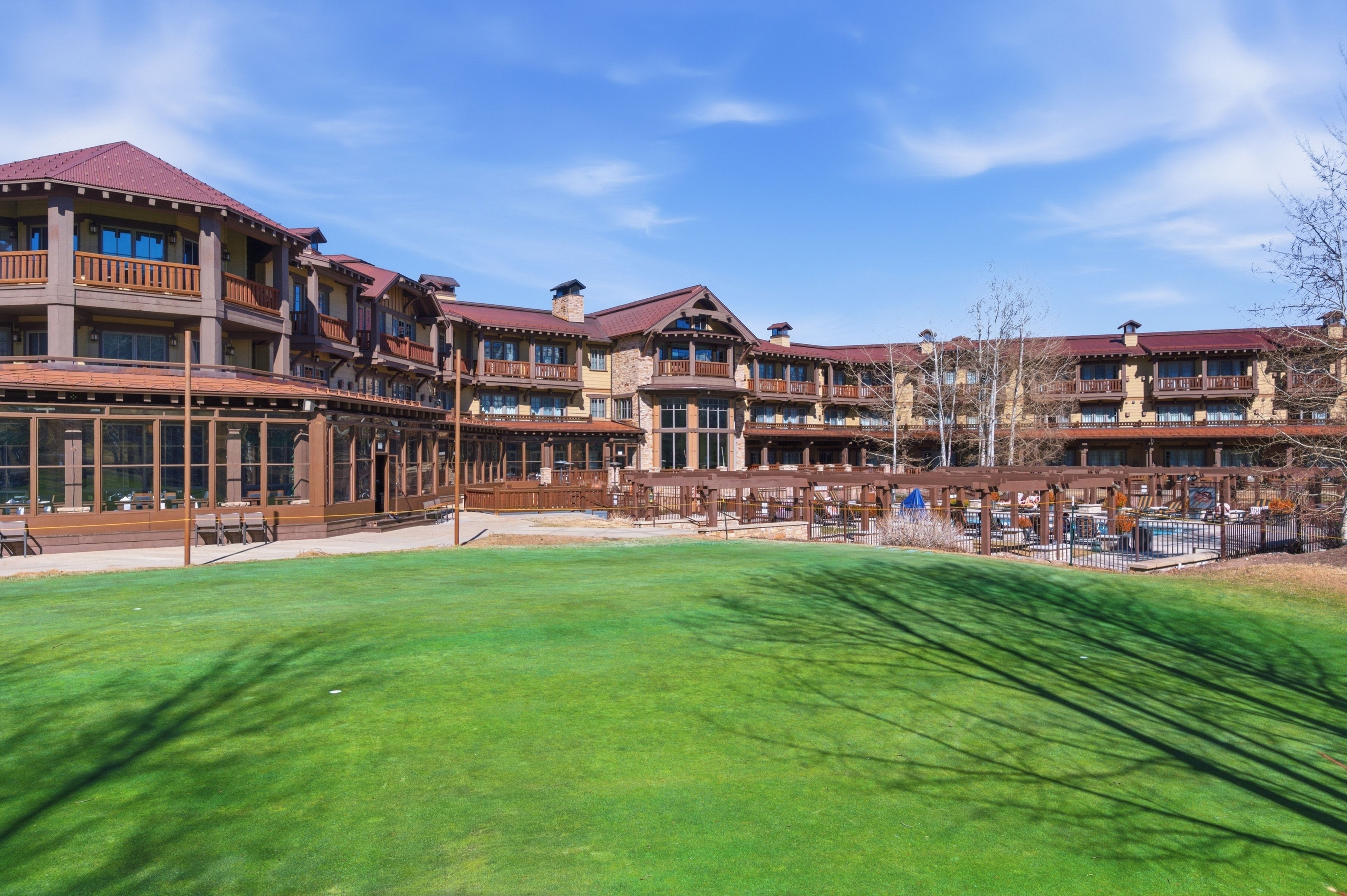 Expansive green lawn fronting a multi-story building featuring natural wood siding, stone accents, multiple balconies, and a red tile roof
