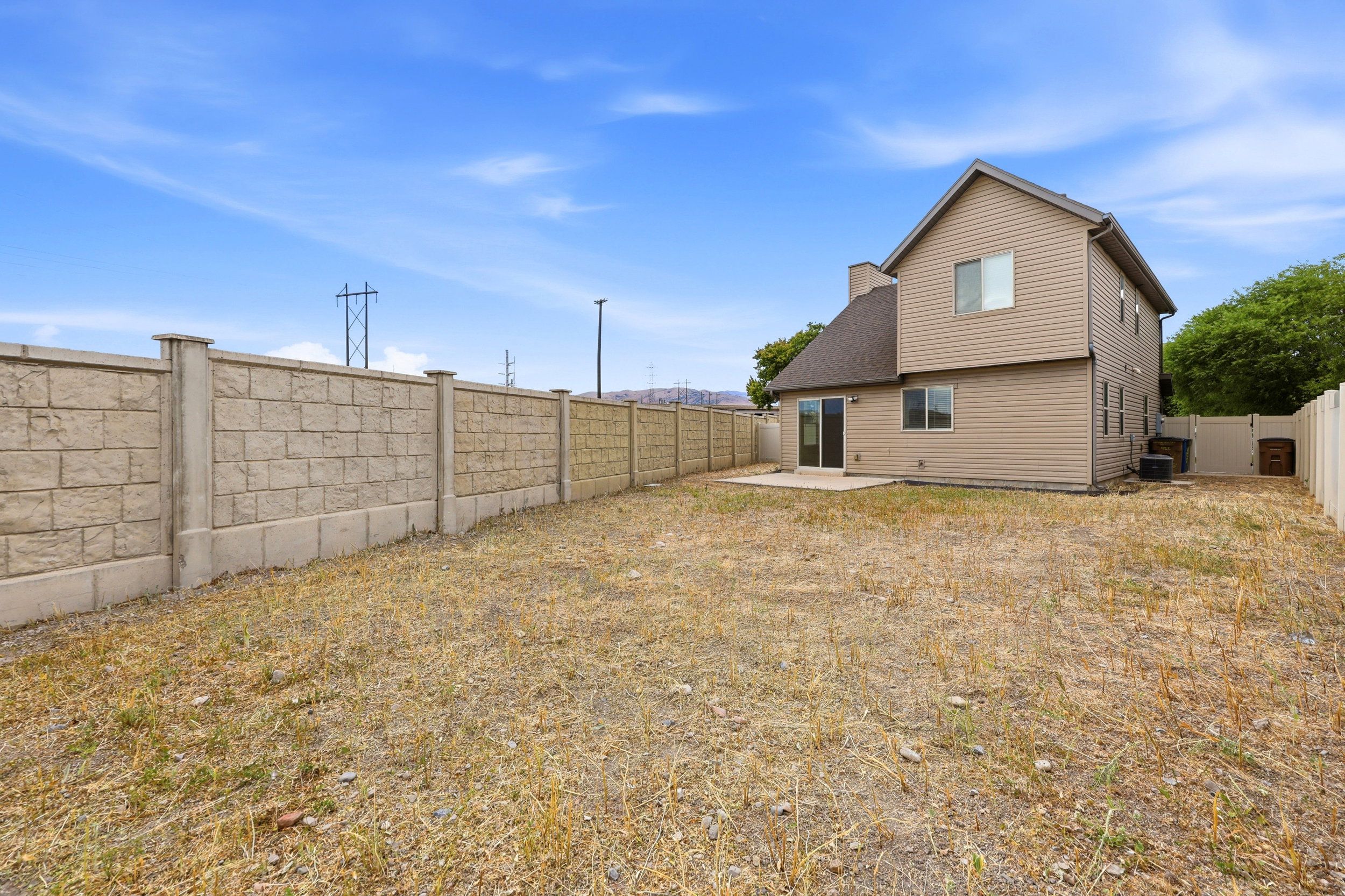 Expansive backyard featuring a concrete block perimeter fence, a tan vinyl-sided exterior, a sliding glass door, and a concrete patio