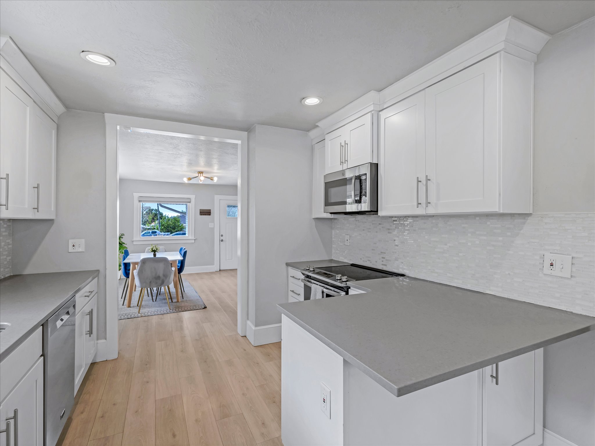 Contemporary kitchen featuring light wood-finish flooring, white shaker-style cabinetry, stainless steel appliances, grey countertops, and a white tile backsplash