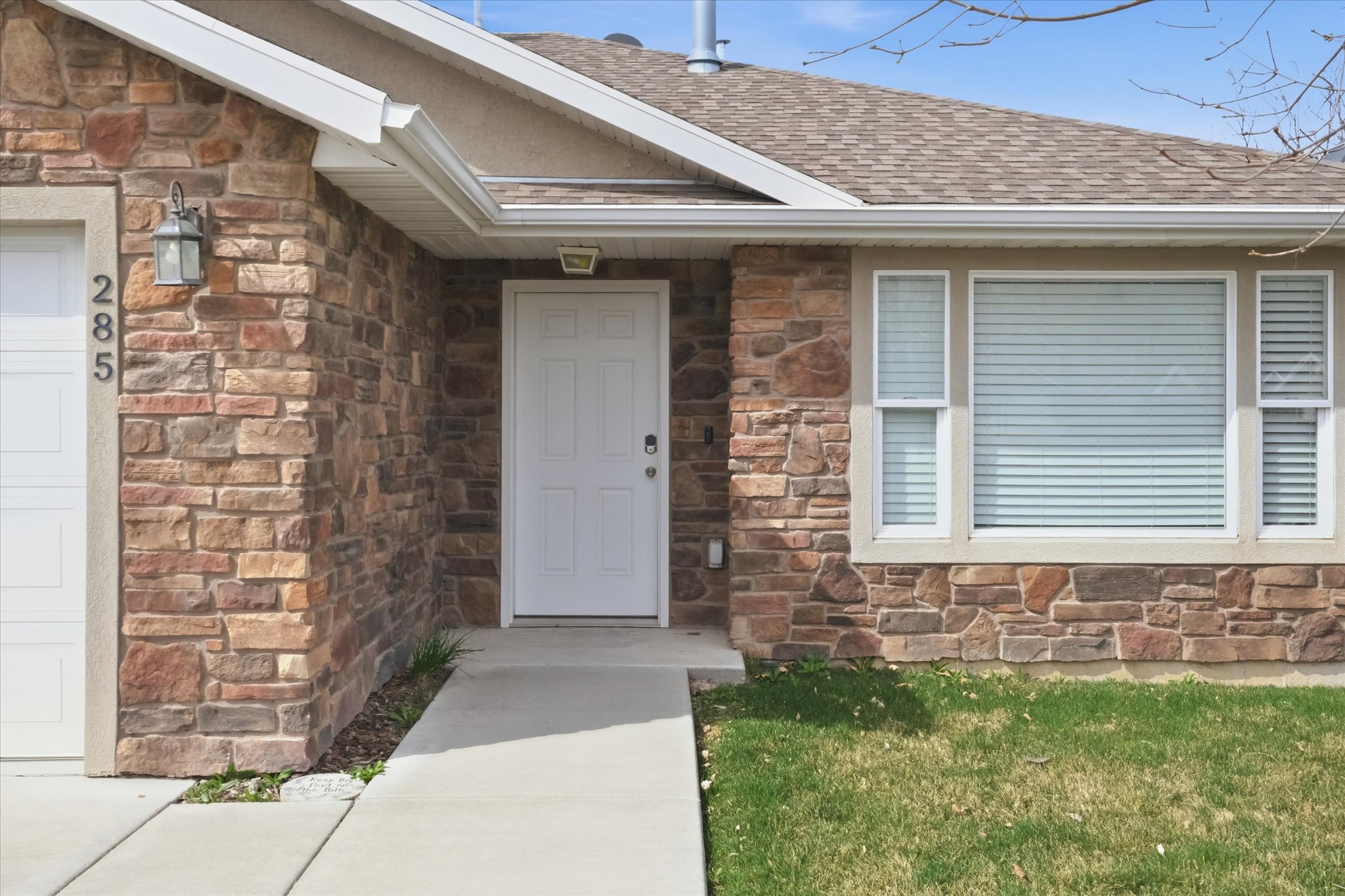 Stone veneer exterior featuring a white paneled entry door, a prominent window with horizontal blinds, and a shingle roof