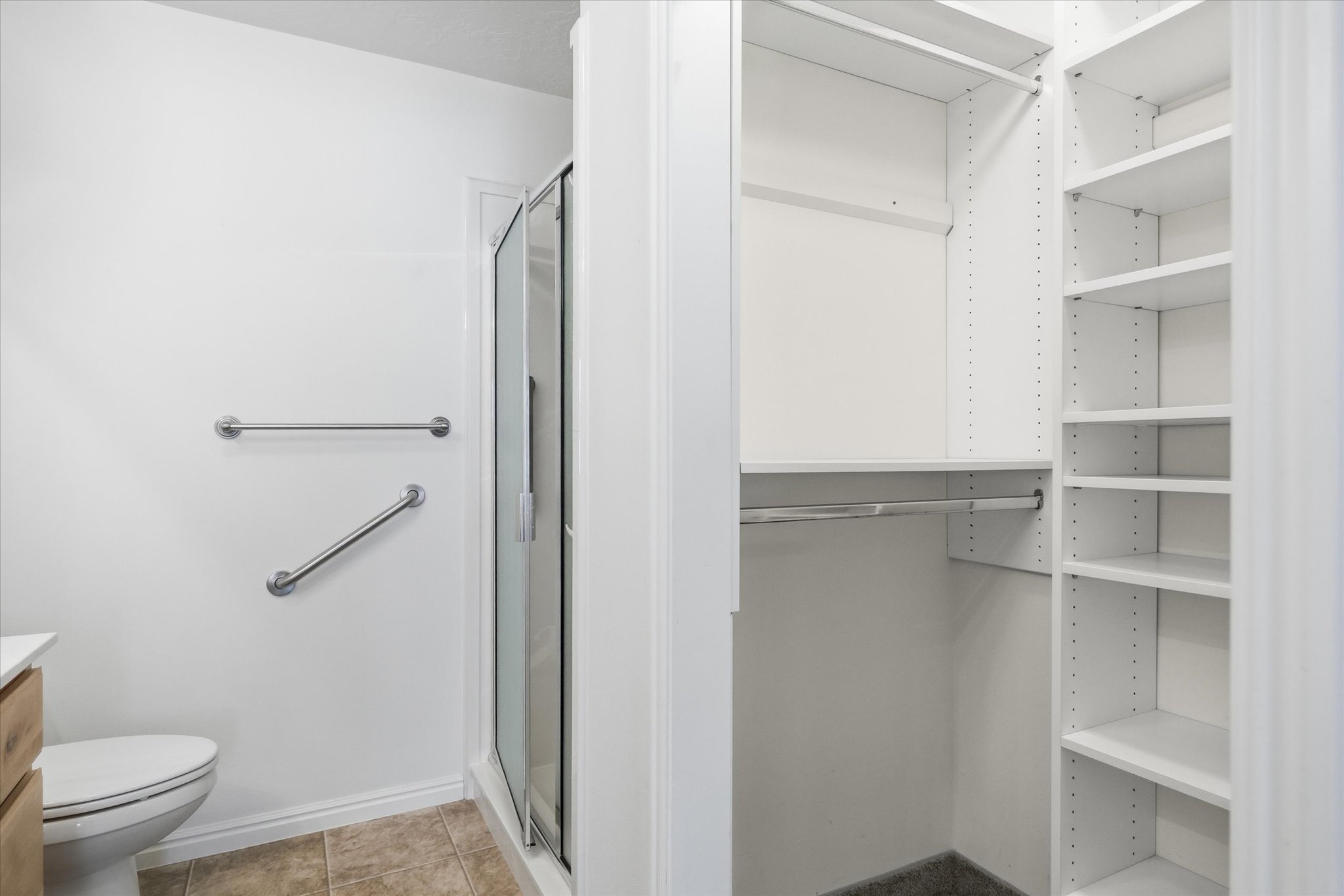 Integrated storage solution featuring a walk-in closet with hanging rods and adjustable shelving, alongside a compact bathroom with a glass-enclosed shower, toilet, and wood-finish vanity