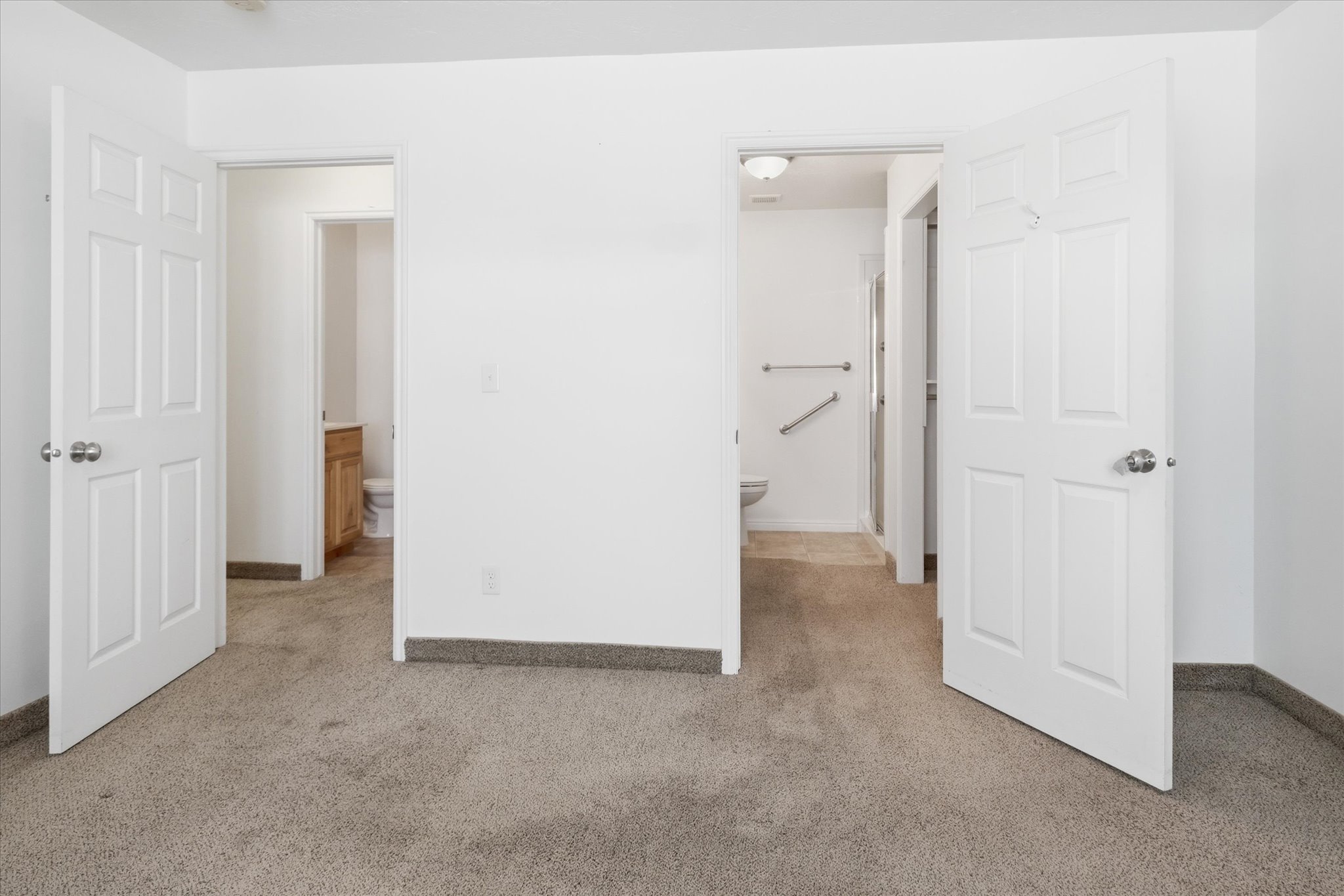 Carpeted interior space featuring white paneled doors with chrome hardware, white walls, and a visible threshold leading into a bathroom with a light wood-finish vanity and toilet