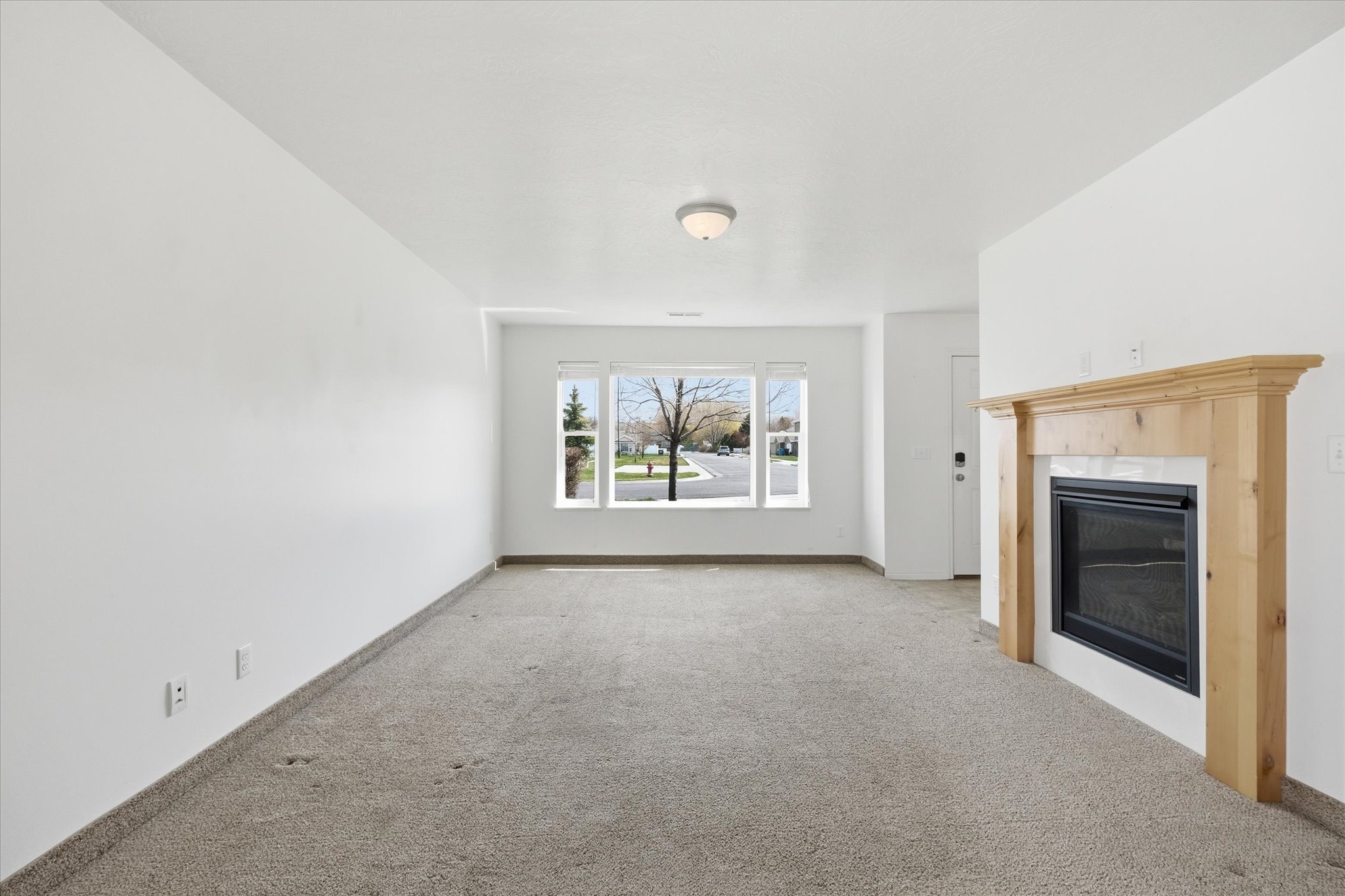Living area featuring a natural wood mantelpiece, a dark-paneled fireplace insert with a white tile surround, carpeted flooring, and a large multi-pane window with white trim