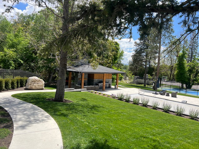 Expansive outdoor area featuring a manicured lawn, a paved pathway, and a covered patio structure with exposed wood beams and stone accents