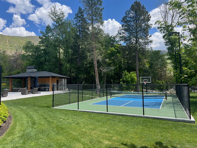 Outdoor recreation area featuring a fenced pickleball court with a basketball hoop, a stone and wood pavilion with a built-in fireplace, and a concrete patio
