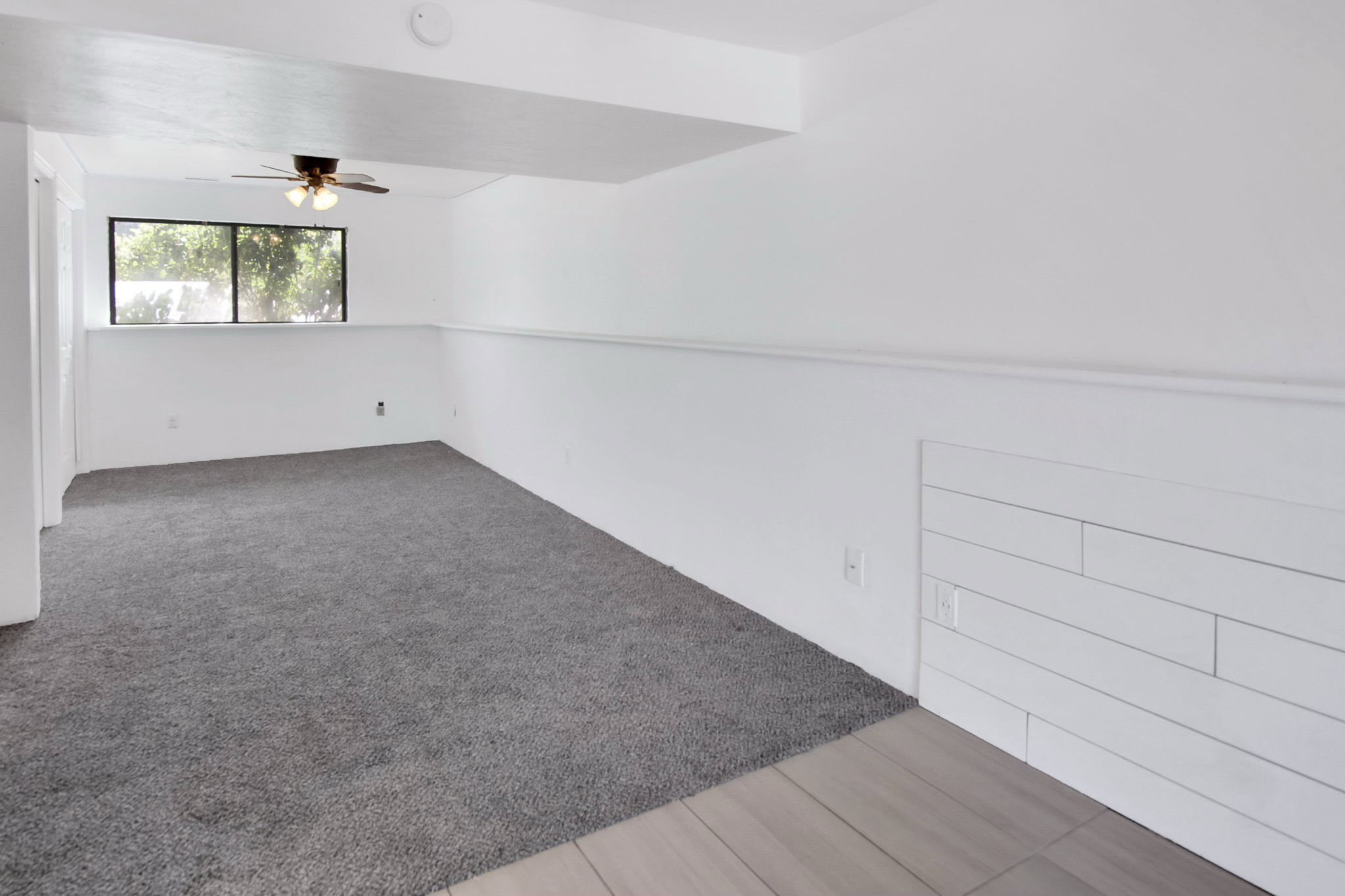 Spacious room featuring a ceiling fan, a large window, gray carpet, and white shiplap wall accents