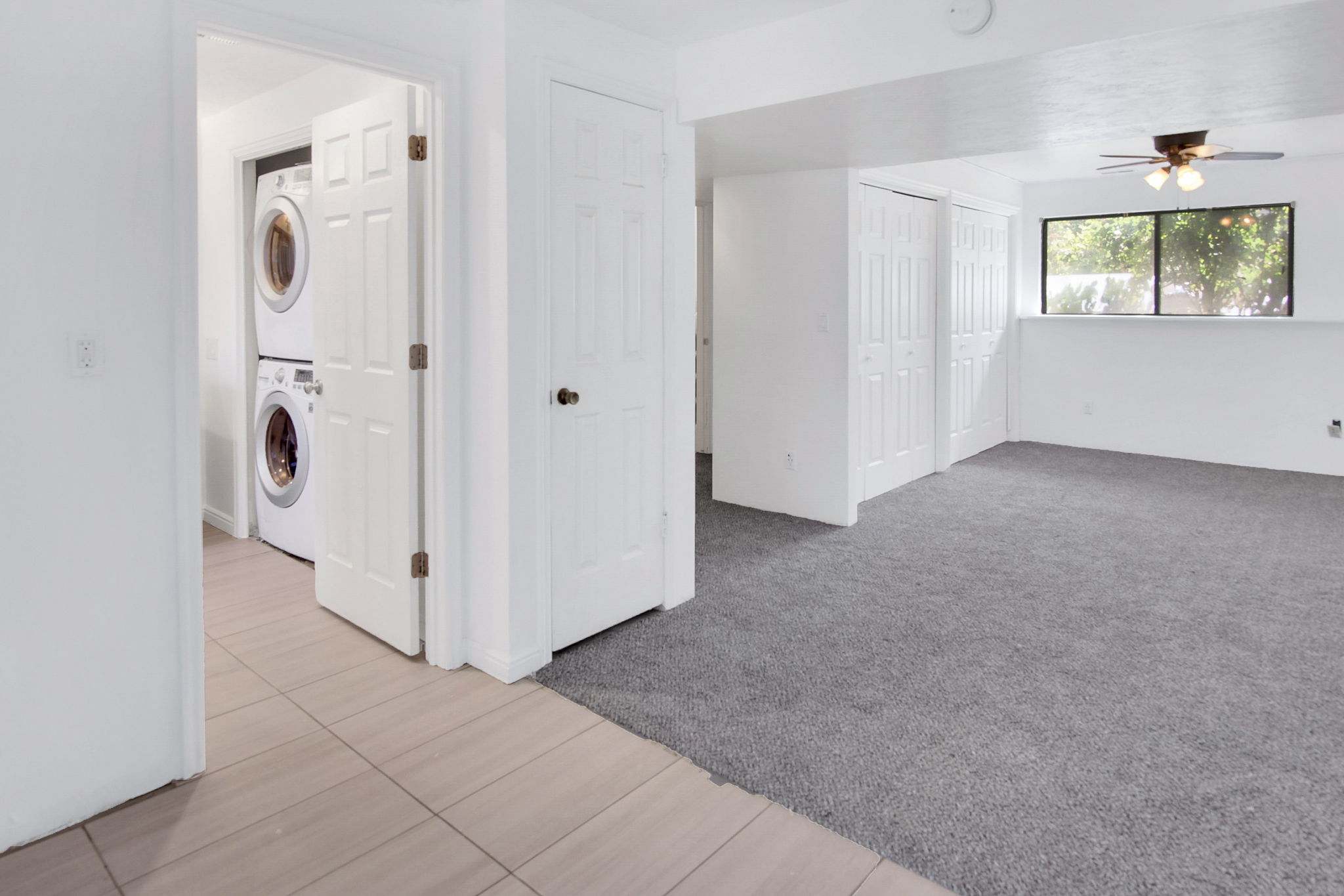 Dedicated laundry area with stacked appliances, complemented by a versatile room featuring grey carpeting, a multi-pane window, and a ceiling fan