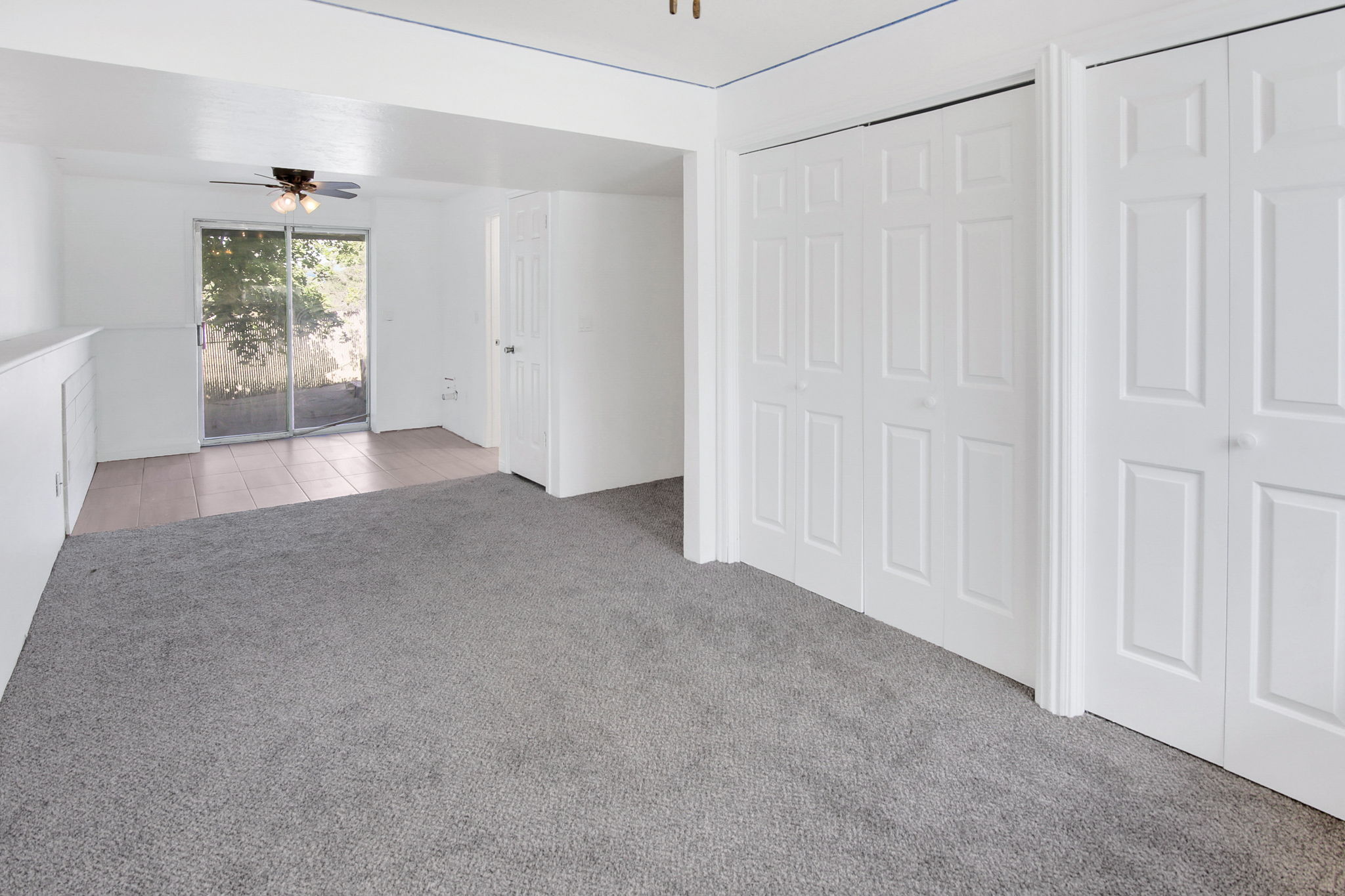 Carpeted interior space featuring white paneled bi-fold doors, a built-in half-wall storage unit, and a sliding glass door with a ceiling fan