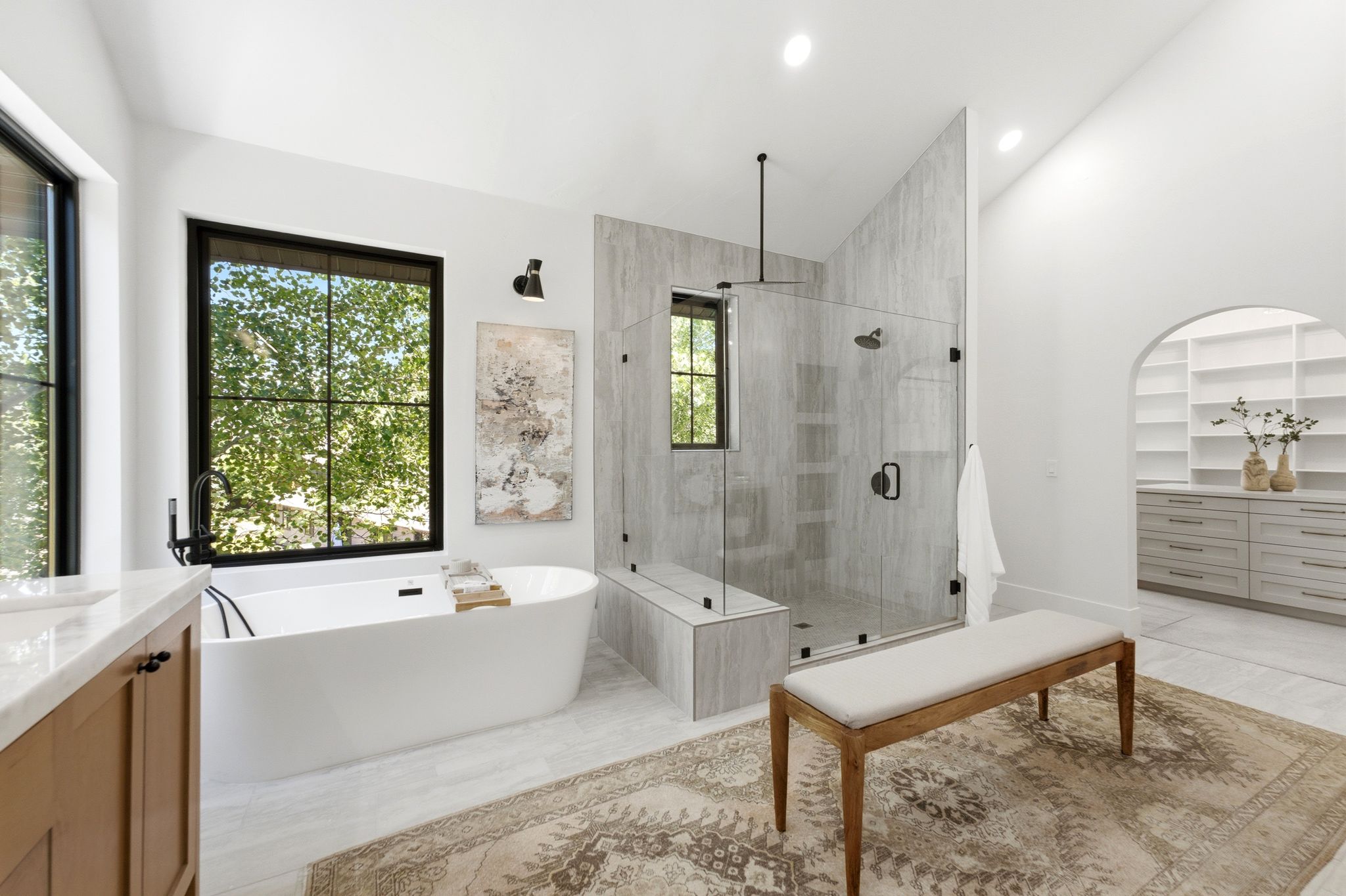 Modern bathroom featuring a freestanding soaking tub with black fixtures, a spacious walk-in shower with a glass enclosure and built-in bench, a large window with black framing, and neutral-toned wall and floor tiling