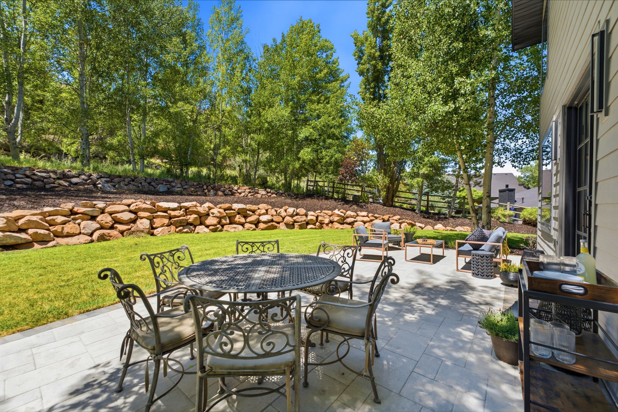 Expansive stone paver patio featuring multiple seating areas, a manicured lawn, and a natural stone retaining wall