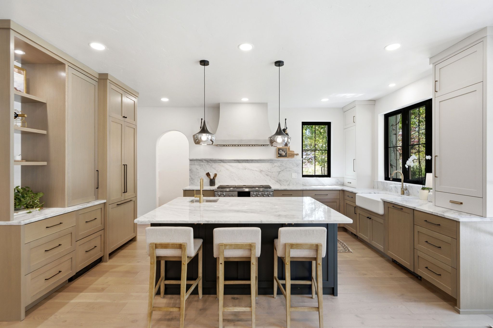 Spacious kitchen featuring a large central island with seating, a farmhouse sink, stainless steel appliances, marble backsplash, and light wood-finish flooring