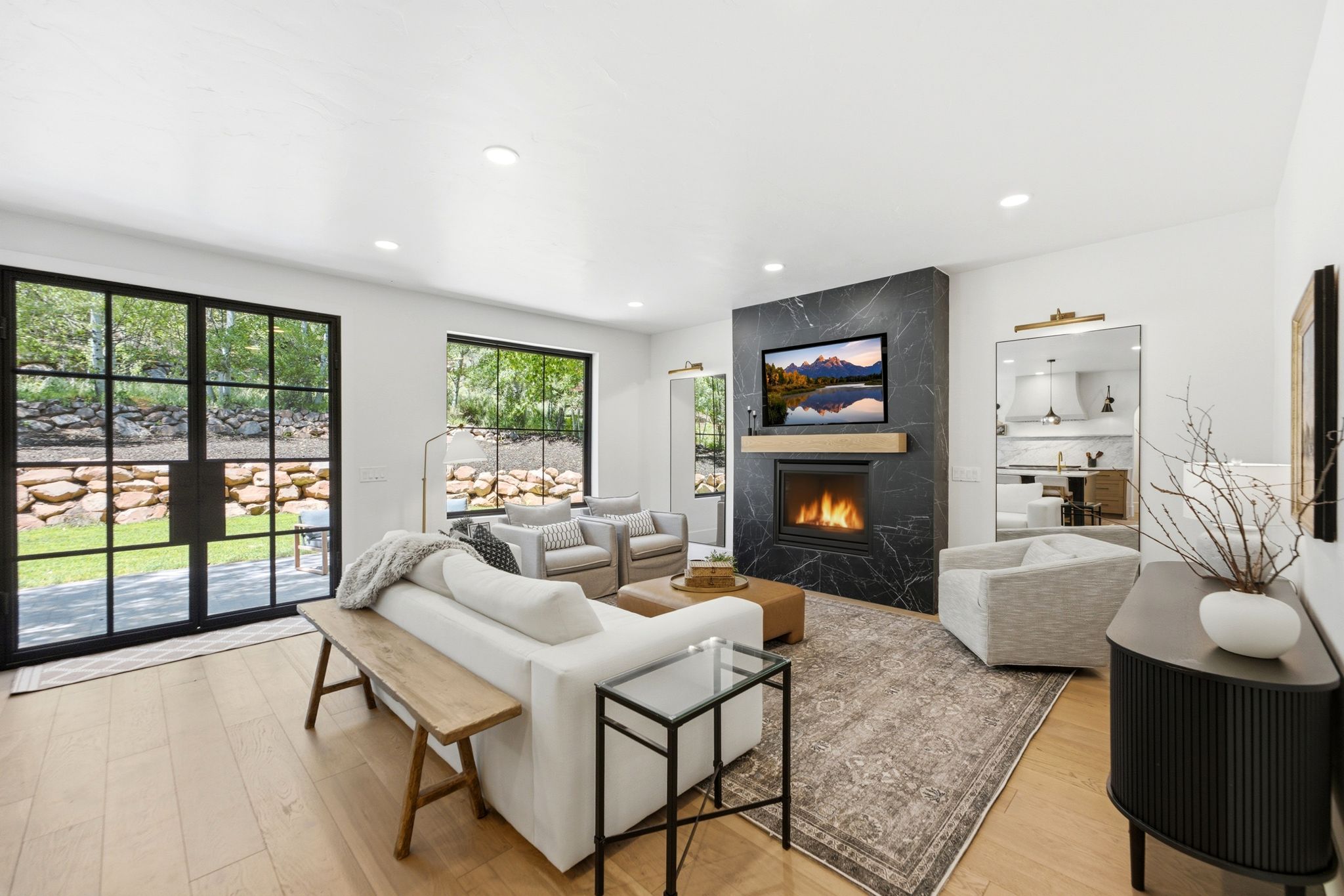 Spacious living area featuring a sleek fireplace with a black stone surround and wood mantel, large black-framed glass doors, light wood-finish flooring, recessed lighting, and a patterned area rug