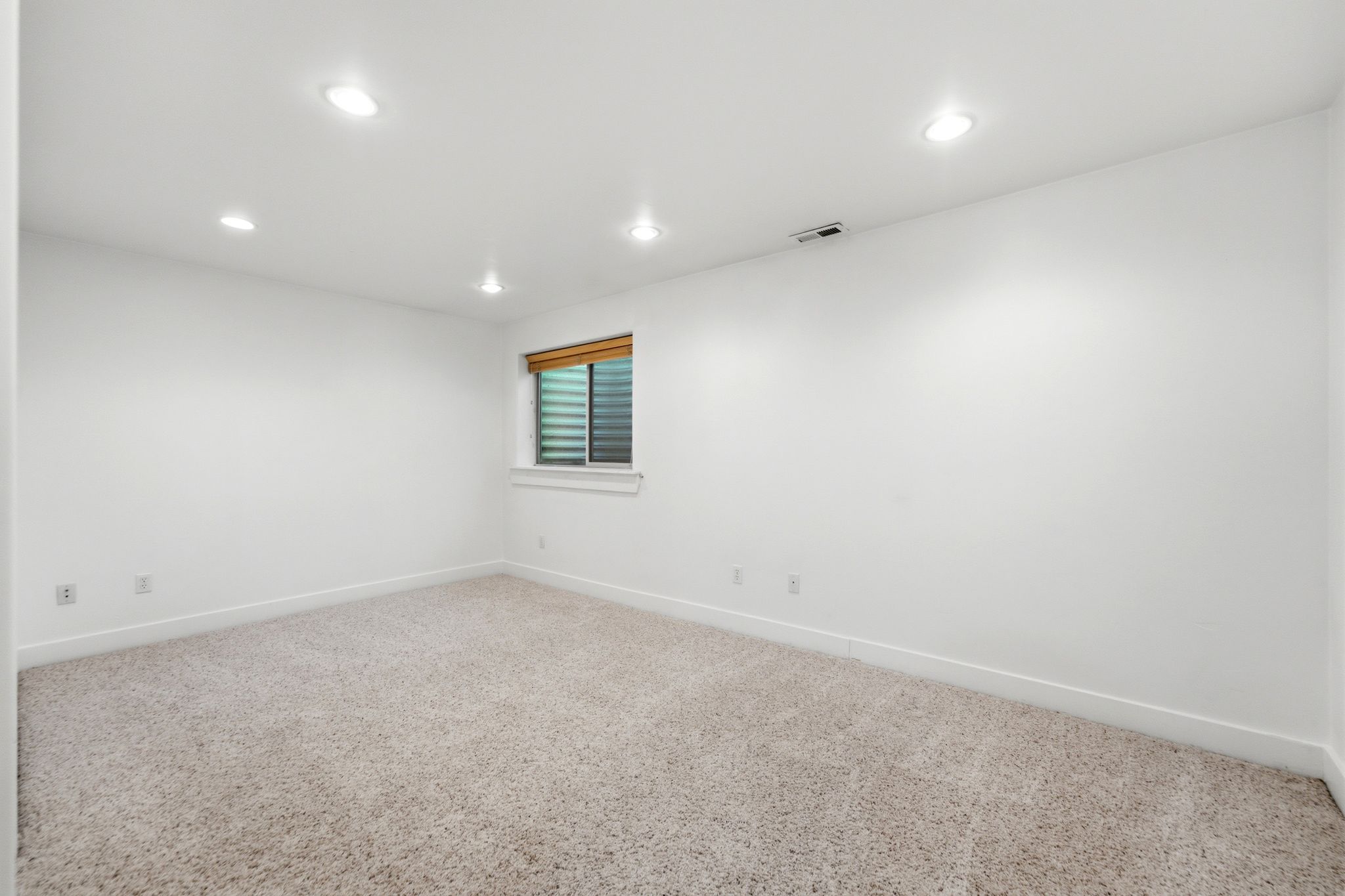 Spacious interior room featuring light-toned carpeting, white walls, recessed ceiling lighting, a window with wood-finish blinds, and white baseboards
