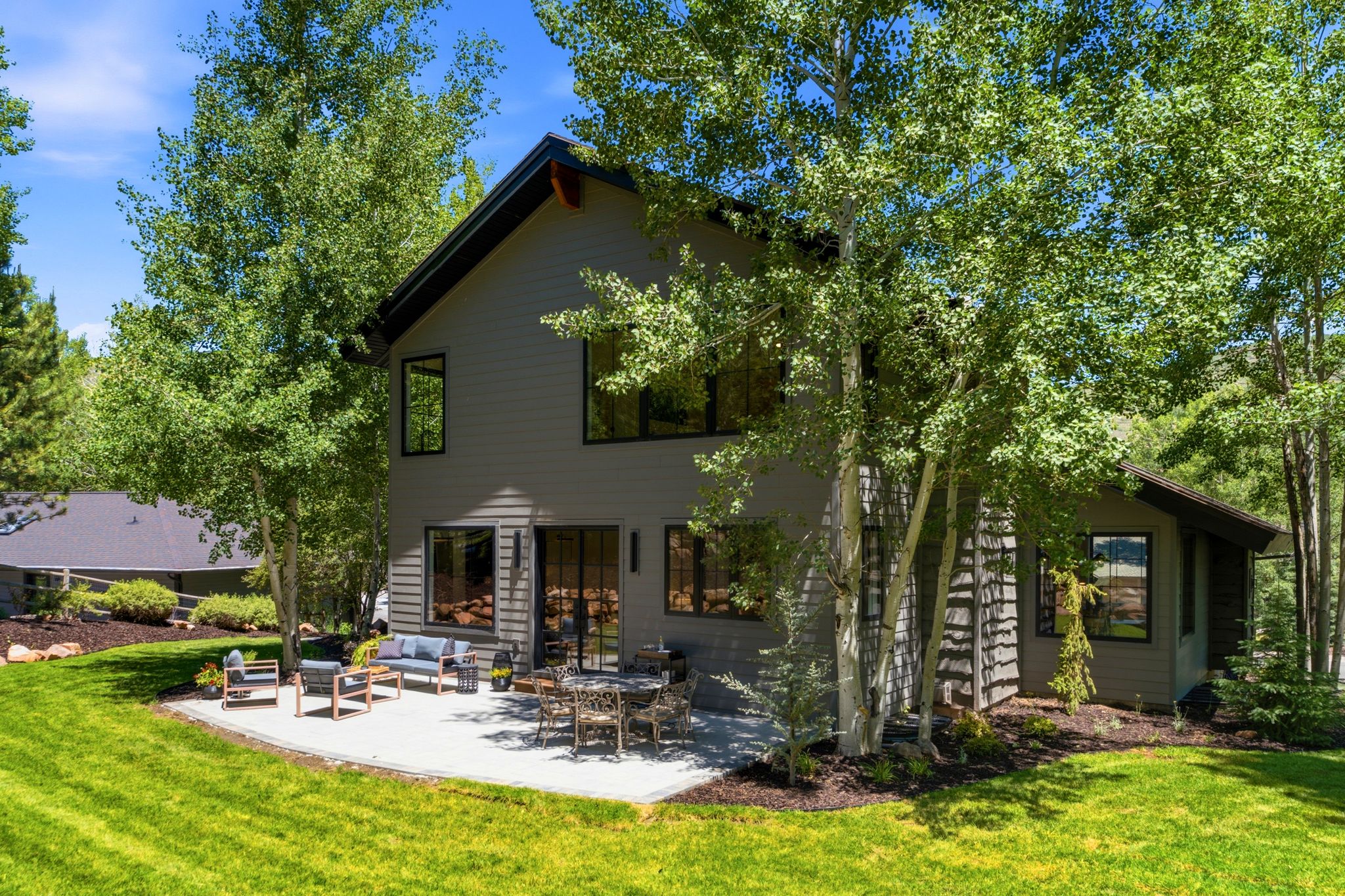 Modern architectural design featuring a spacious concrete patio, a manicured lawn, abundant mature trees, and a dark-toned exterior with large windows and a glass slider