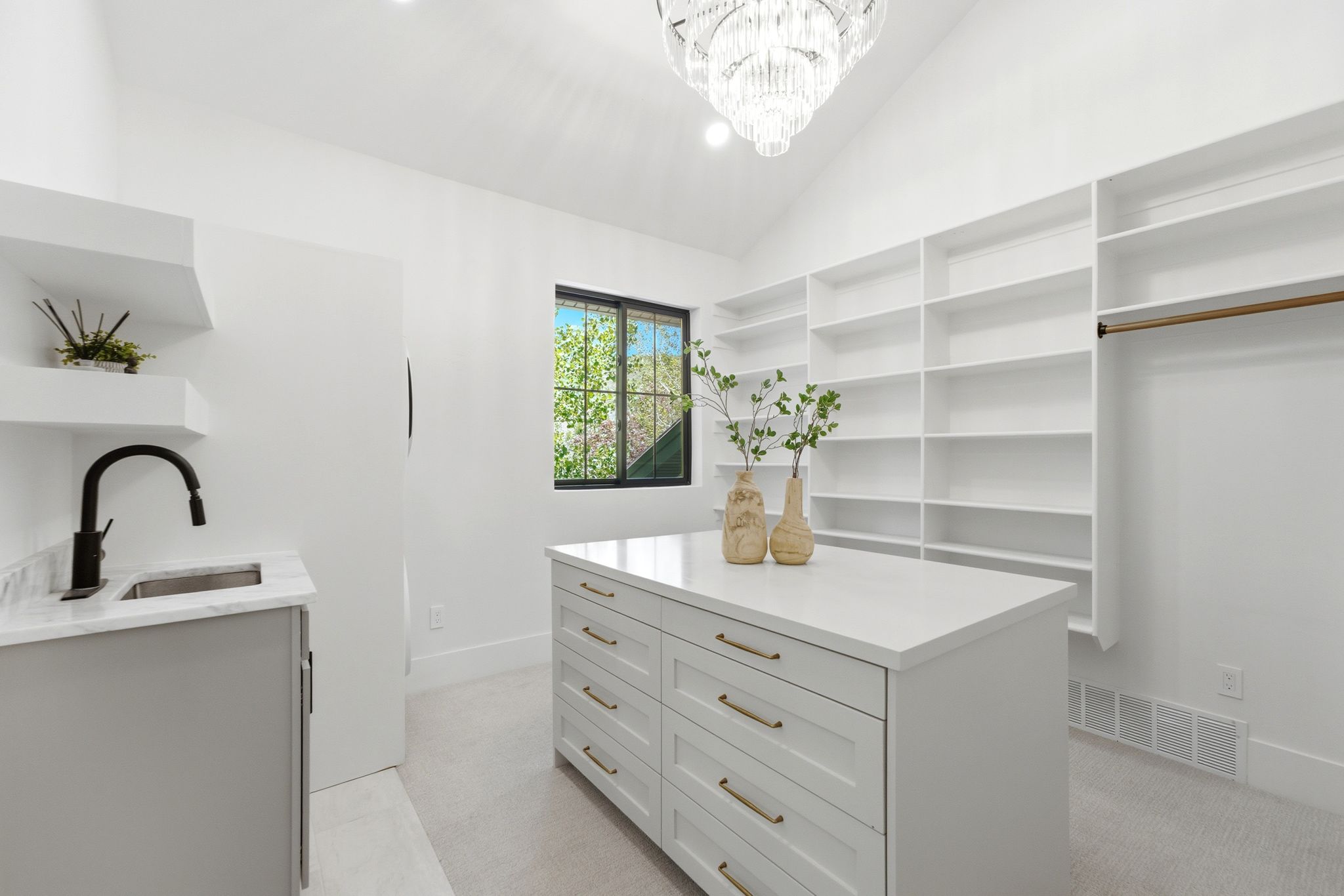 Vaulted ceilings with a chandelier illuminate this space featuring a built-in island with storage, extensive built-in shelving, a dedicated sink area with floating shelves, and a window for natural light